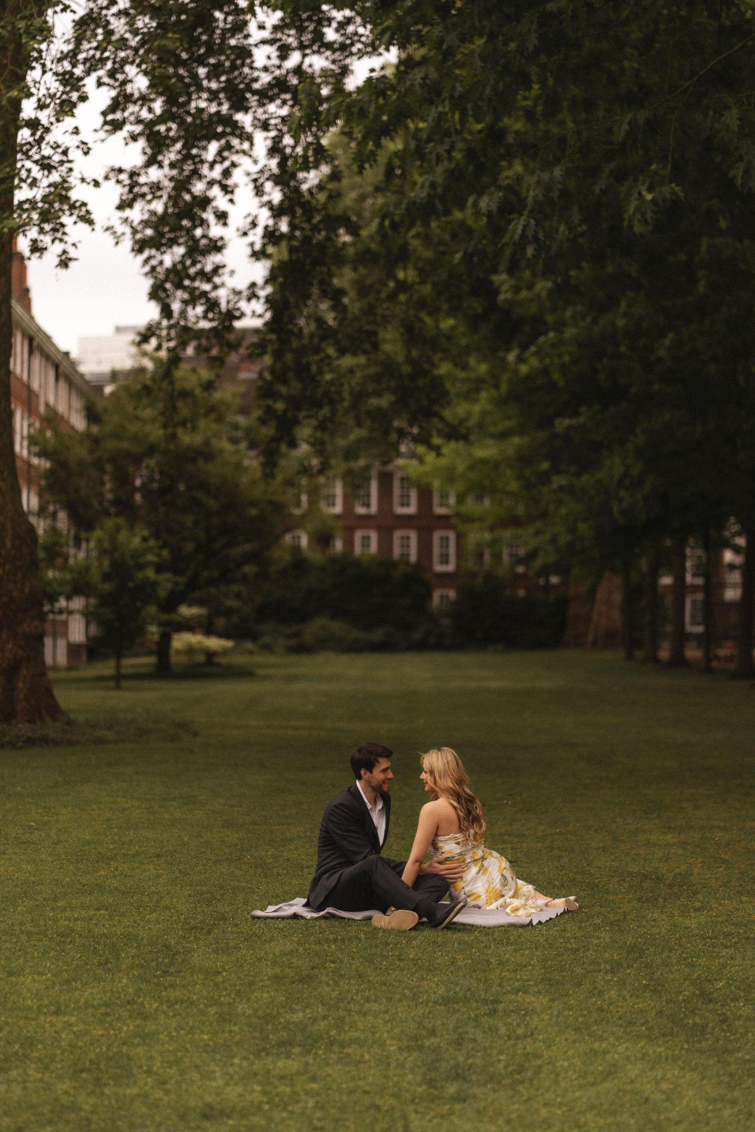 A couple sits together on a lush green lawn in front of historic brick buildings on a summer evening.
