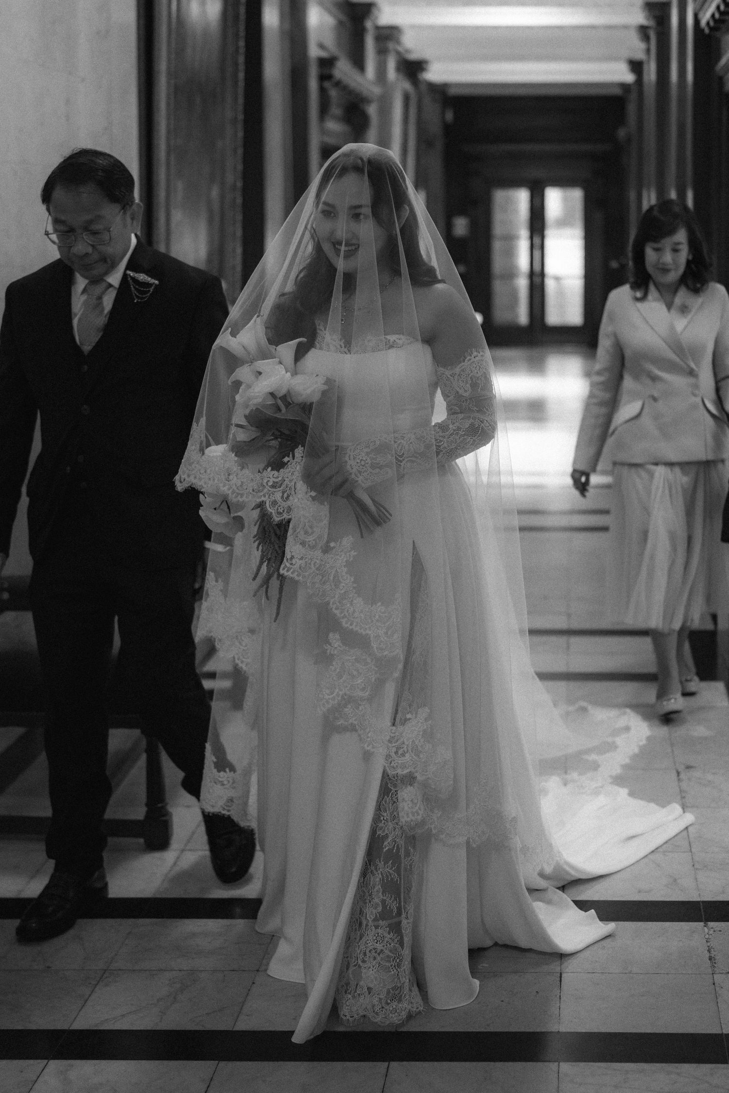A black and white wedding photograph showing a bride in a long veil and dress walking down a hallway with two attendants.