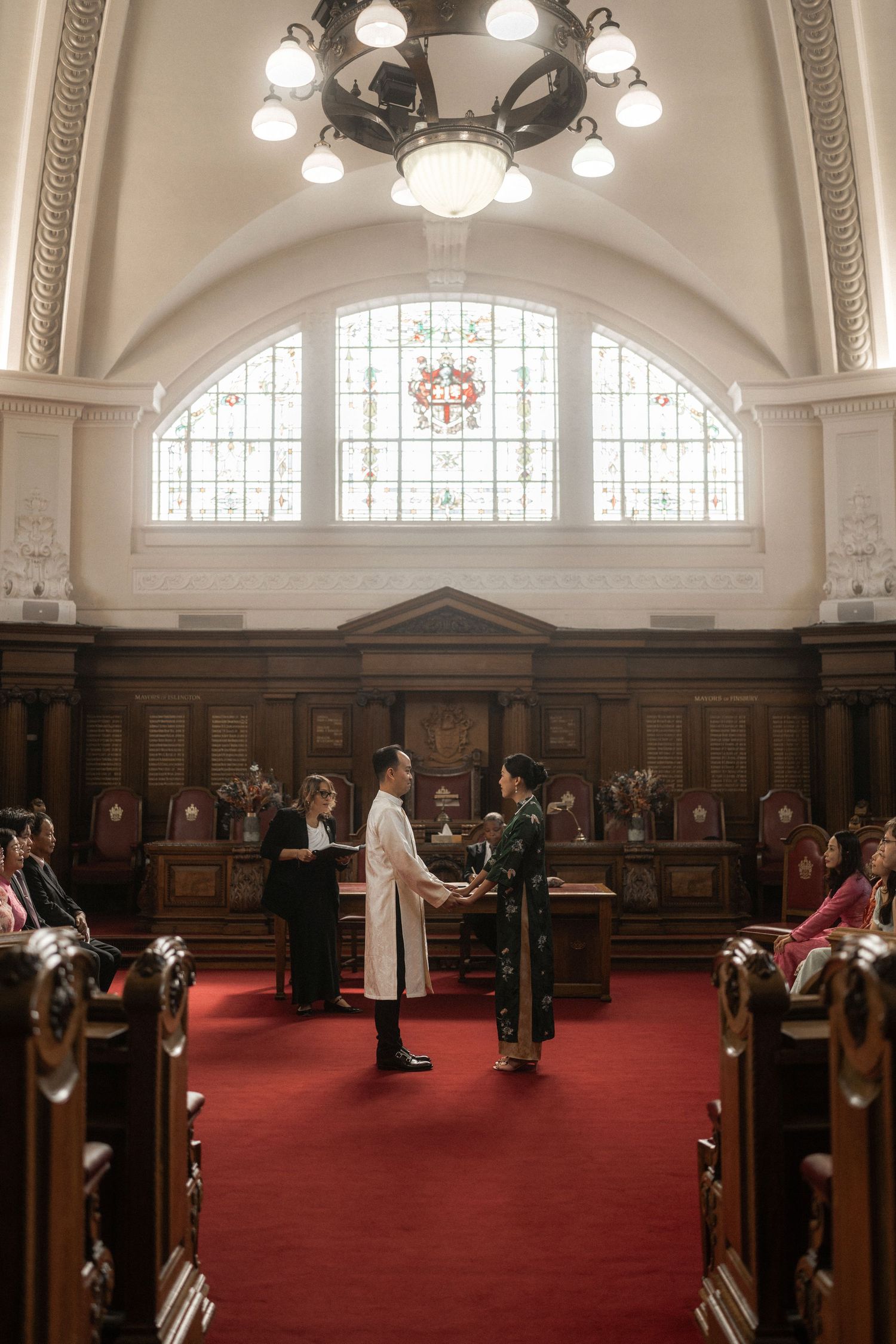 Couple exchanging vows in an elegant wood-paneled ceremony hall with stained glass windows and red carpet aisle.
