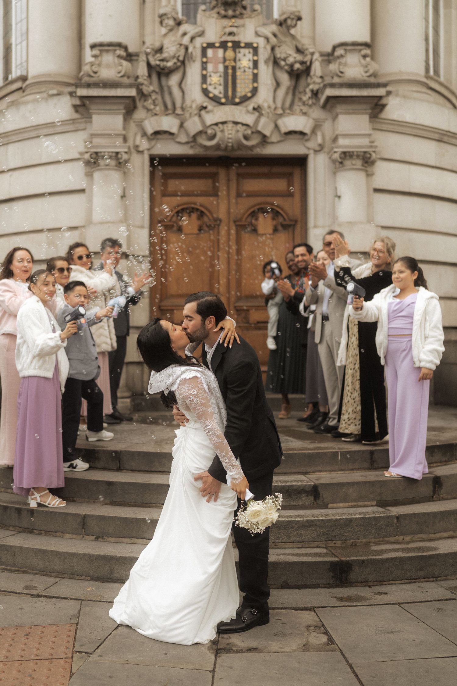 Bride and groom share a kiss on church steps while guests throw confetti at their wedding ceremony.