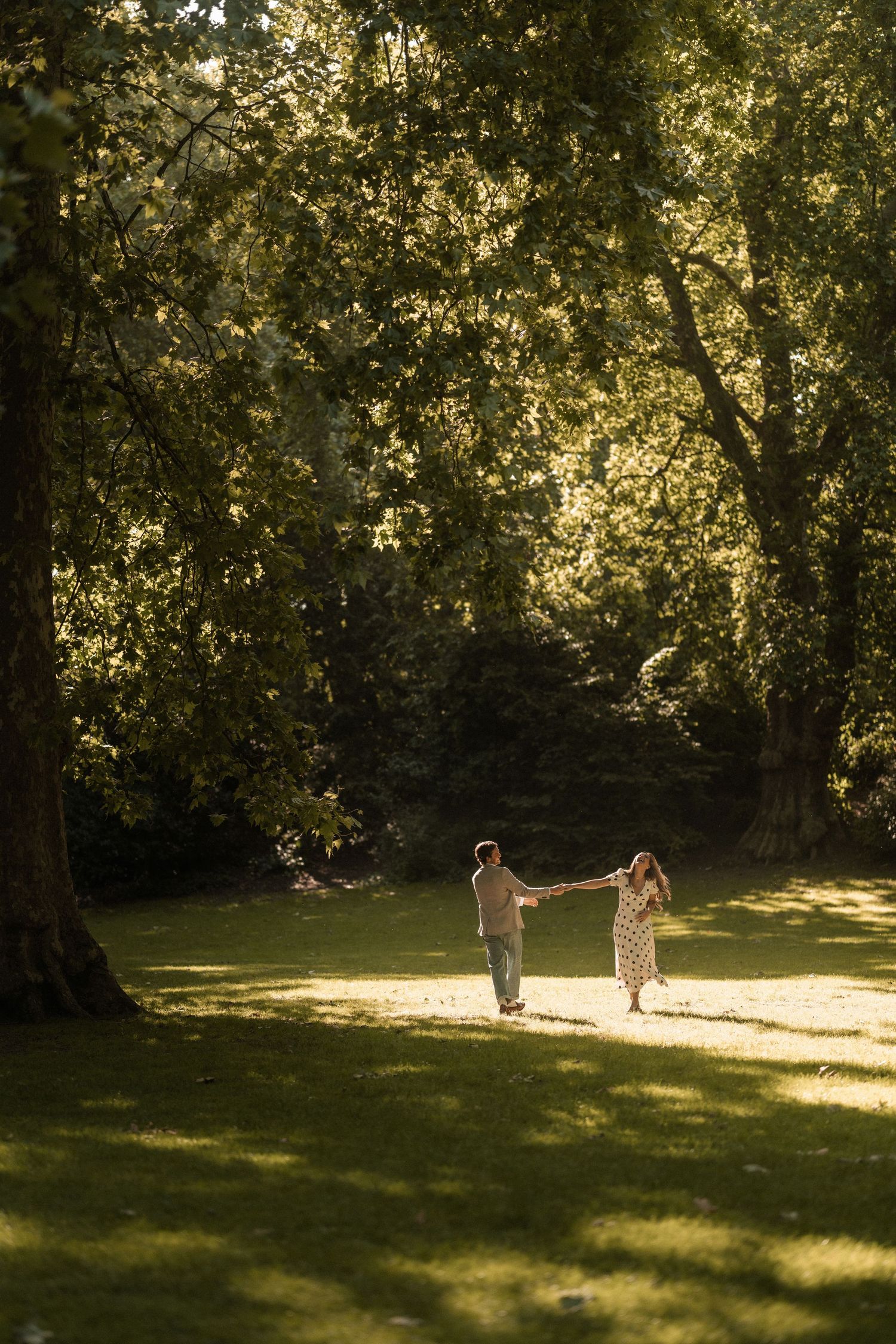 Couple holding hands and walking through a sunlit green park surrounded by tall trees on a warm day.
