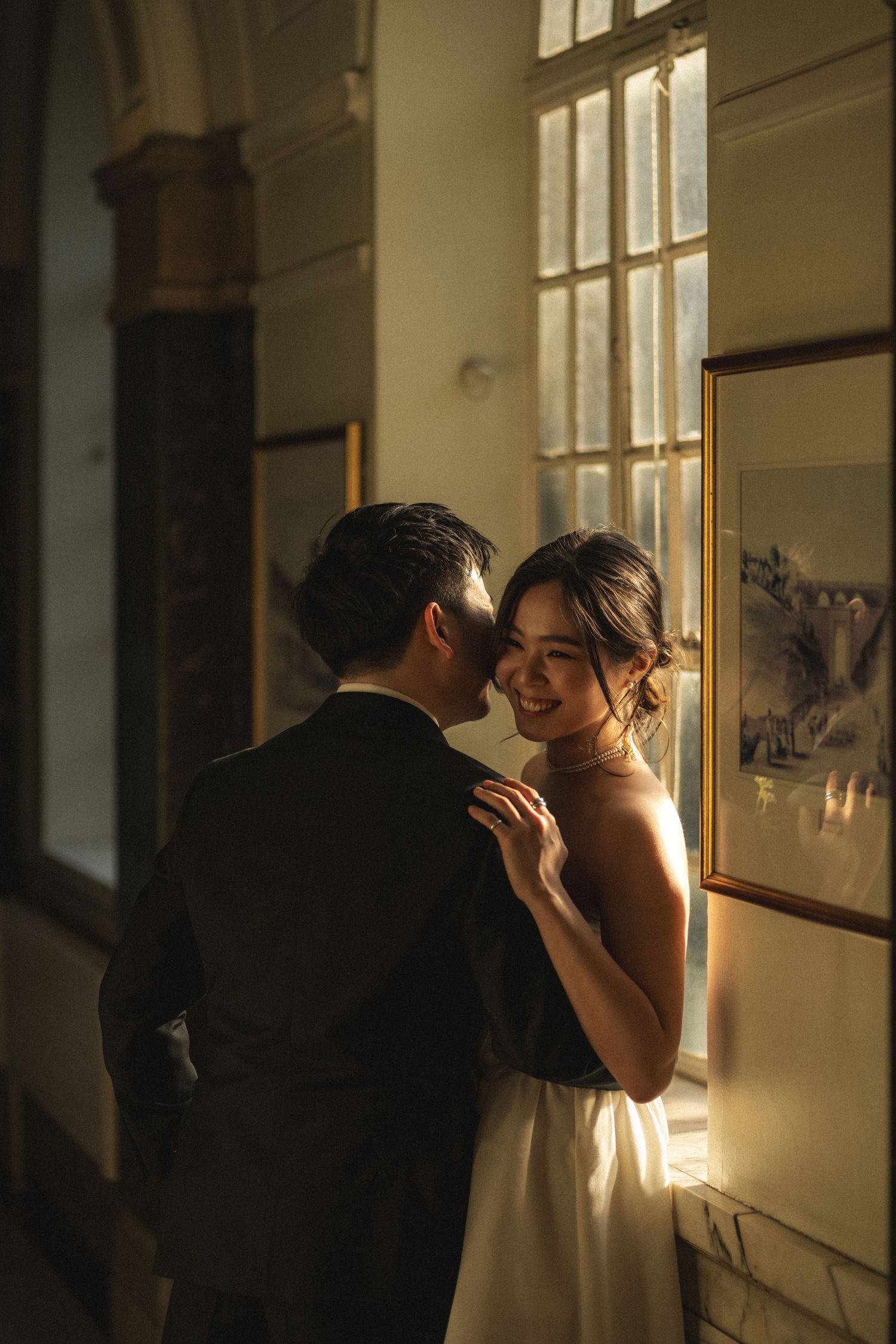 A romantic couple shares an intimate moment by a sunlit window, with the bride wearing an elegant white dress.