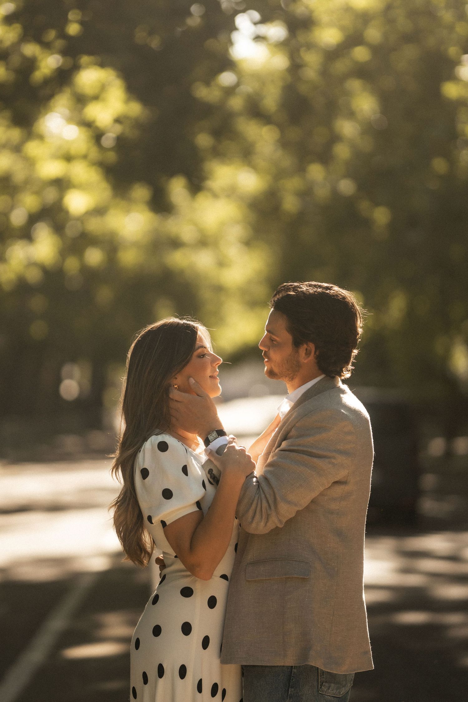 Couple embracing tenderly in a sunlit park, woman in polka dot dress and man in gray blazer gazing lovingly at each other.