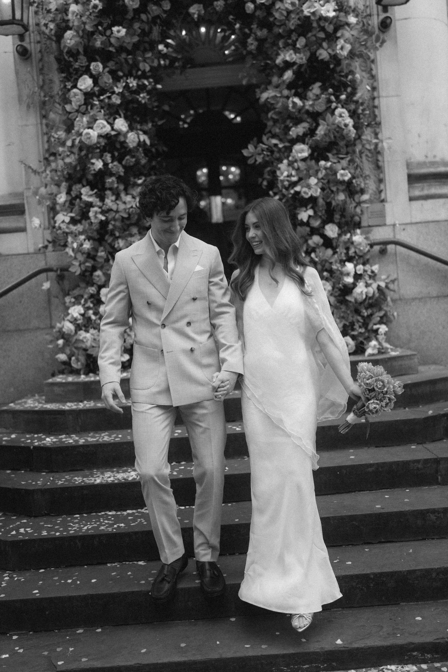Black and white photo of a couple in wedding attire holding hands on steps adorned with floral arch.
