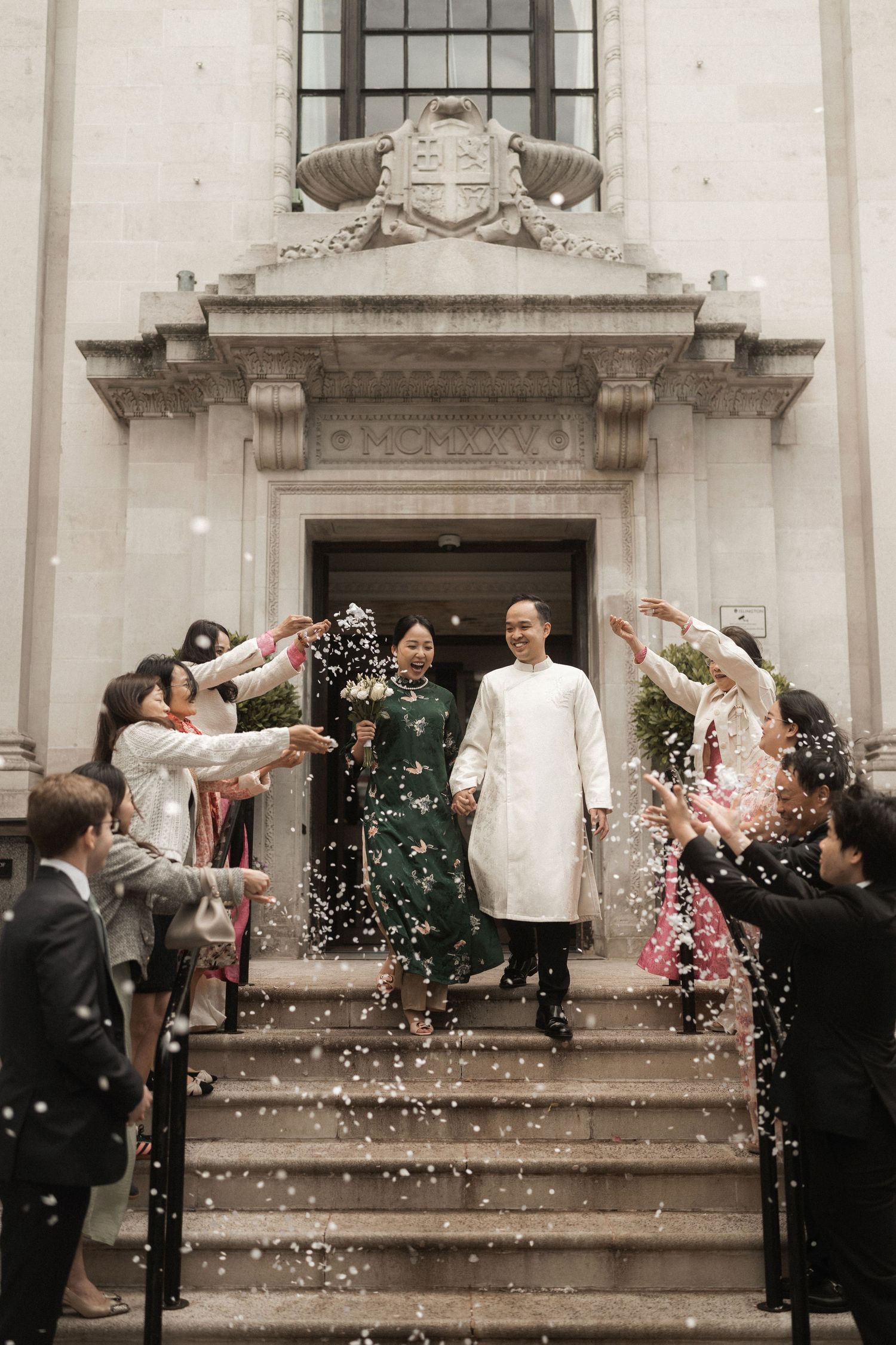 Couple in traditional Vietnamese ao dai exits grand building as guests toss white confetti during wedding celebration.