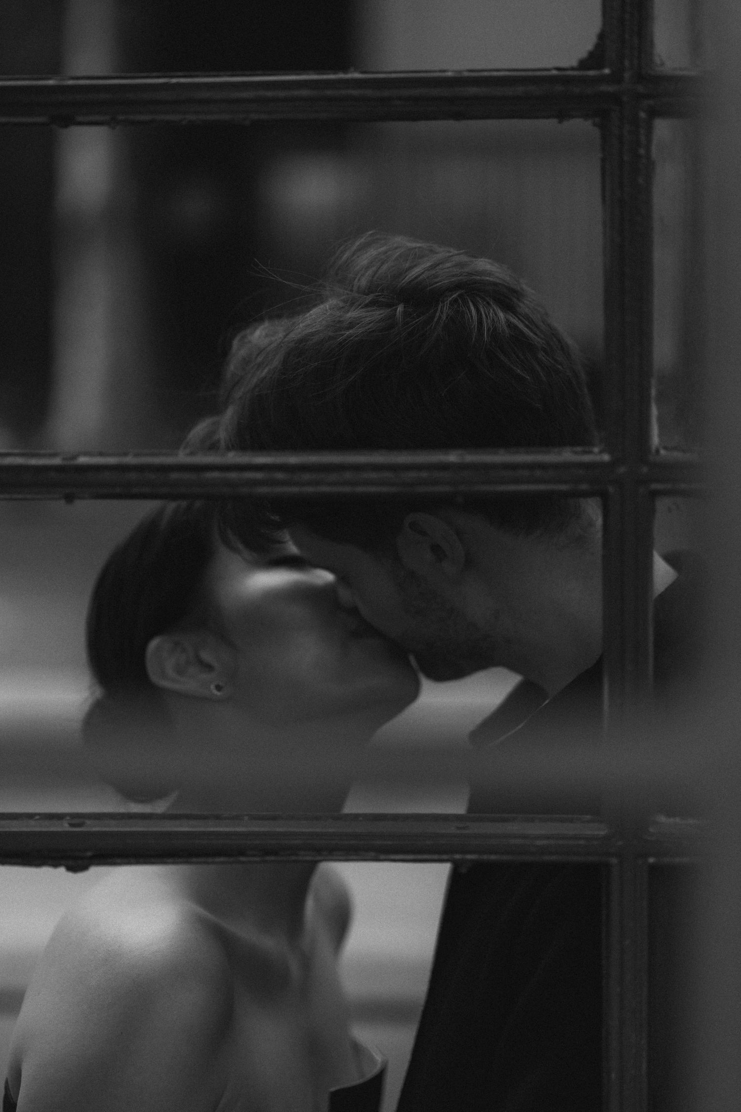A romantic black and white photograph of a couple sharing an intimate kiss through window panes.