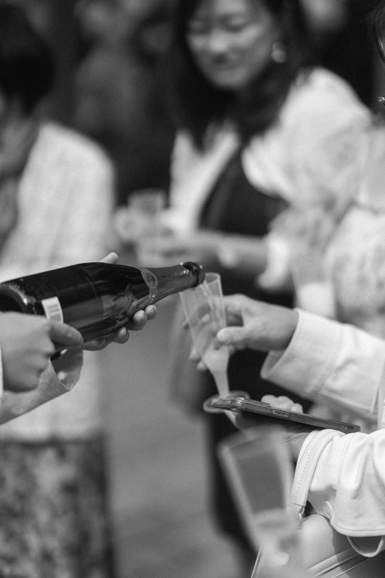 Black and white photo of a champagne bottle being poured into glasses at a social gathering.