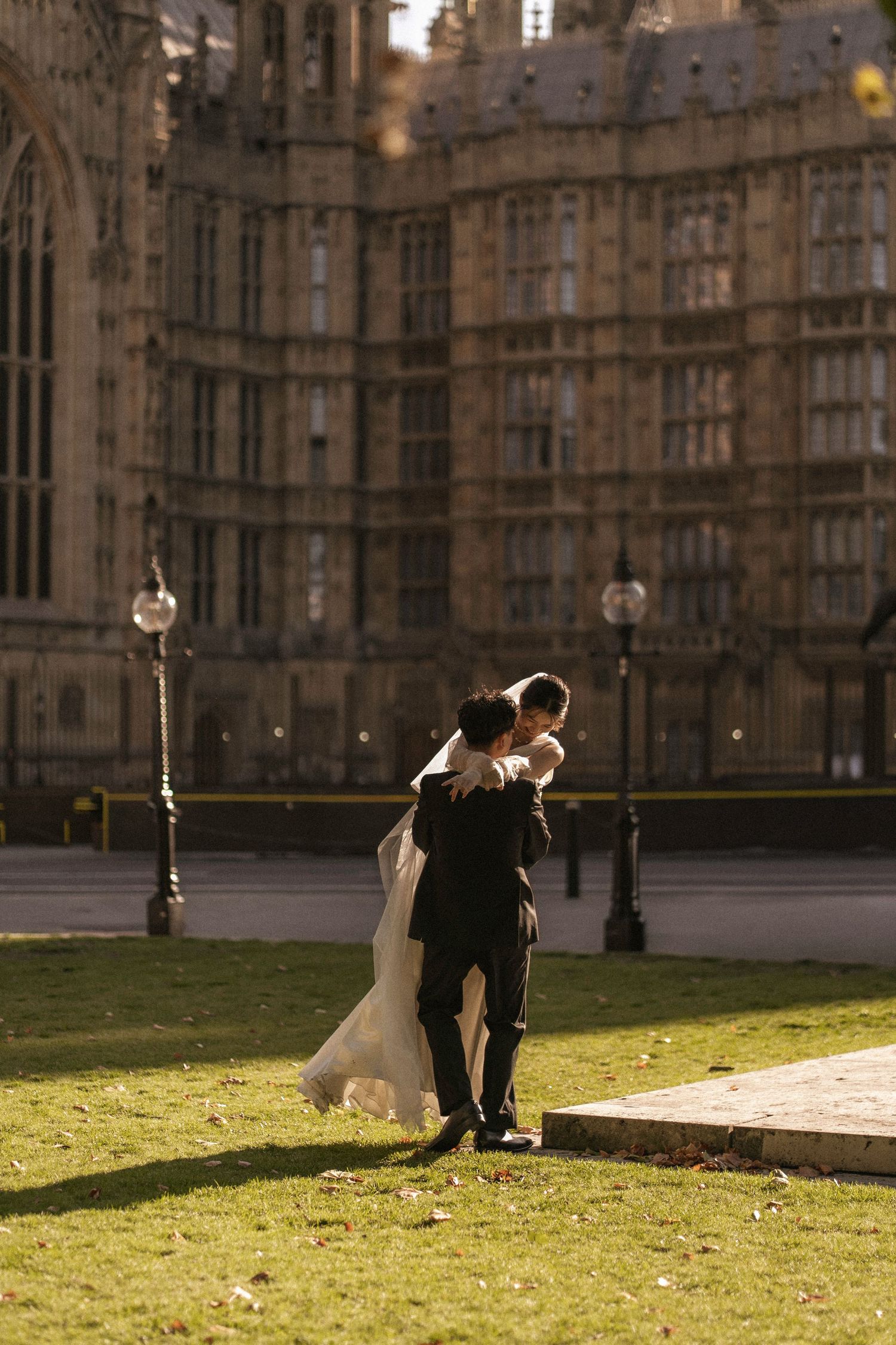 Bride and groom embracing on green lawn in front of the Houses of Parliament, London, on a sunny day.