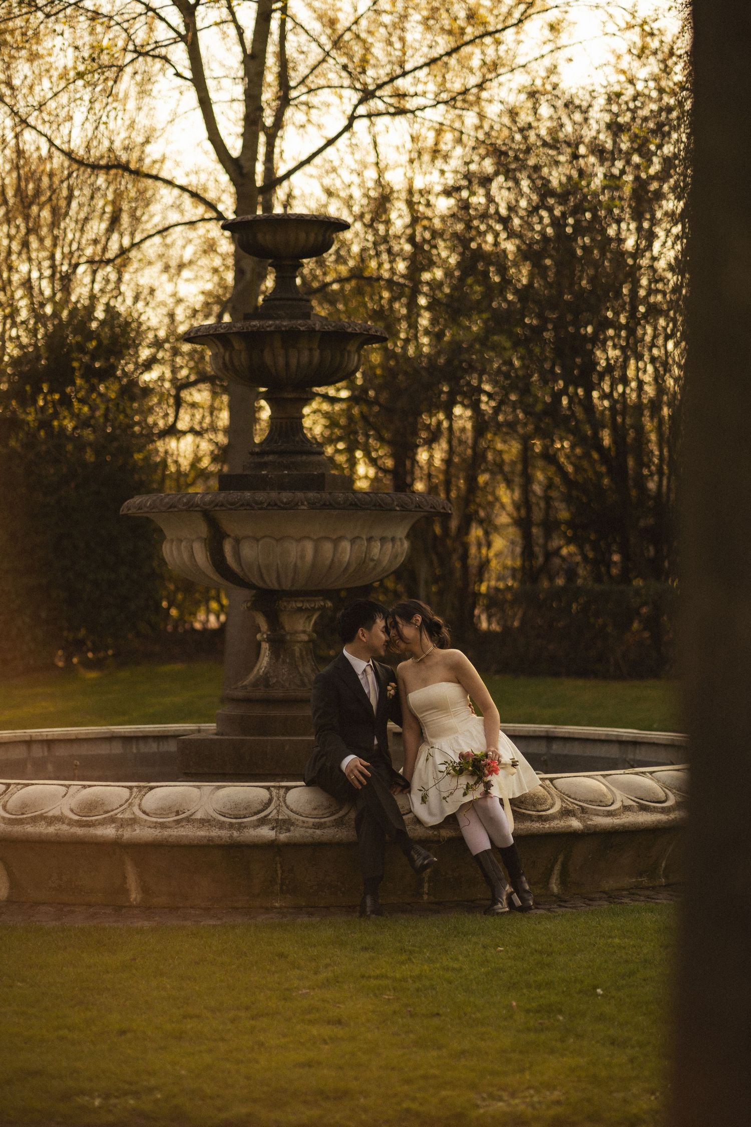 A couple sits intimately by an ornate garden fountain at sunset, surrounded by lush grounds and golden light.