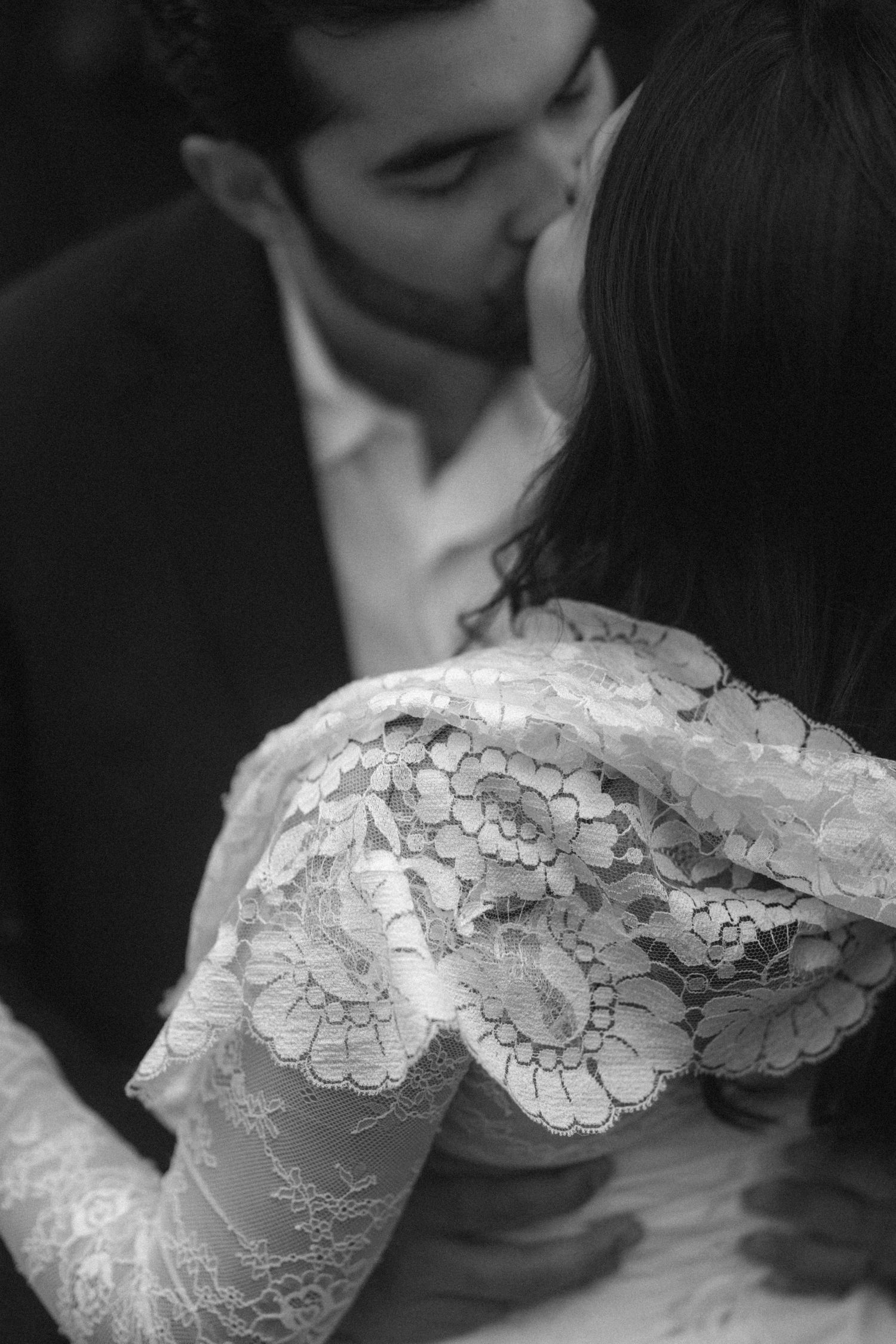 Black and white photo of a couple sharing a romantic kiss, bride in elegant lace dress embracing groom in dark suit.