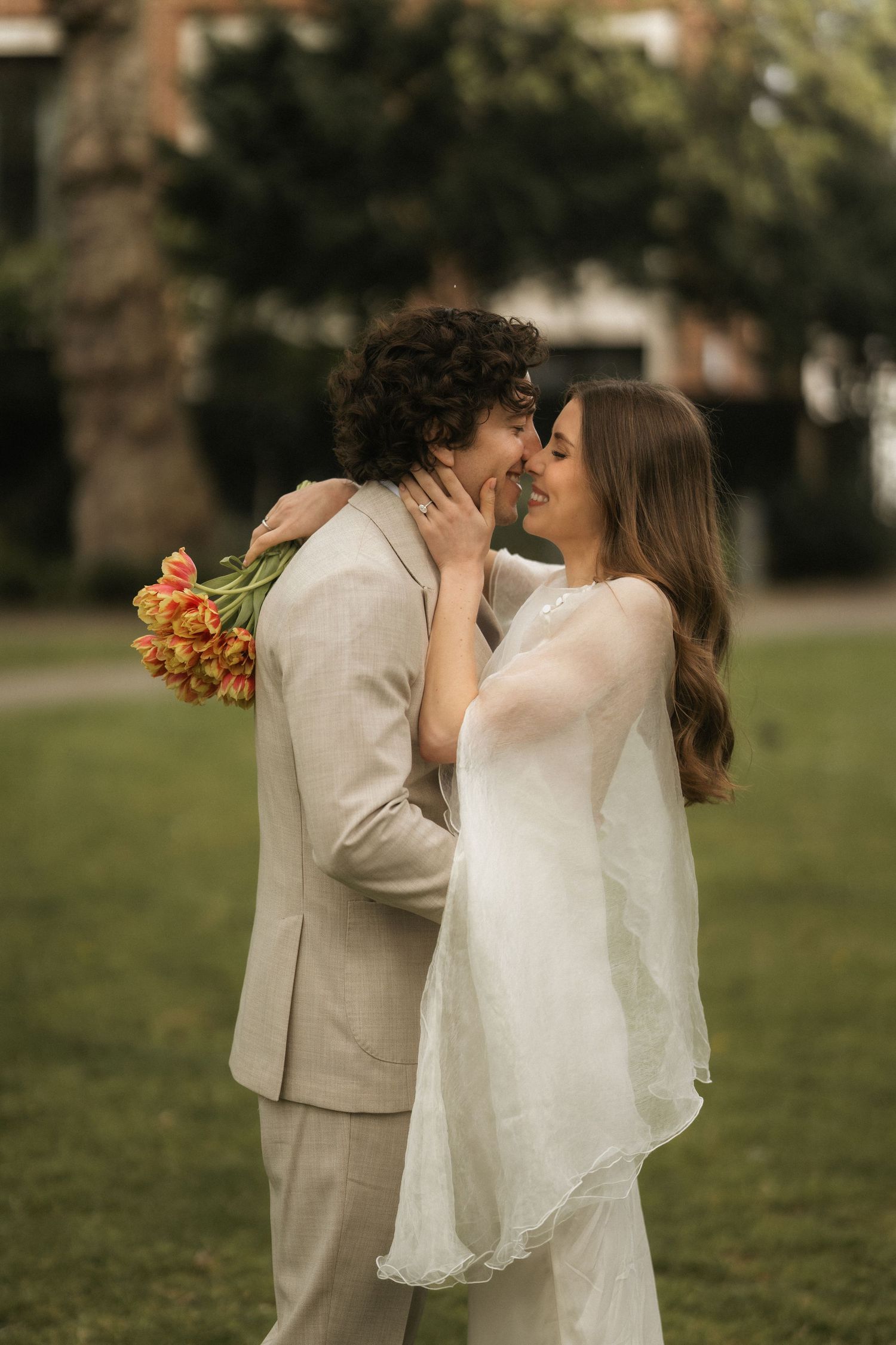 A bride and groom share a romantic kiss outdoors, she in a white gown holding tulips, he in a beige suit.