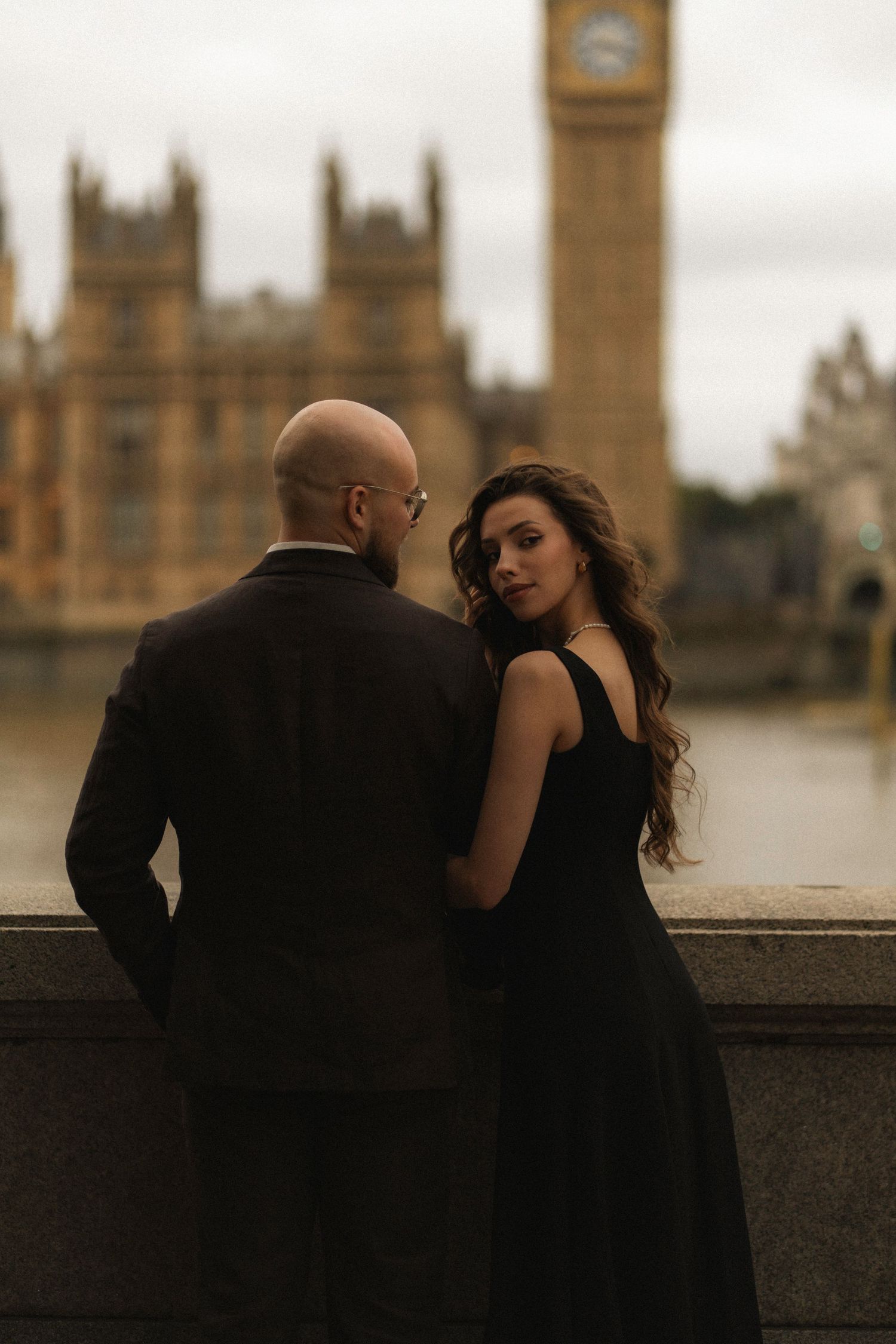 Elegant couple in black attire posing near the Thames with Big Ben and Houses of Parliament in background.