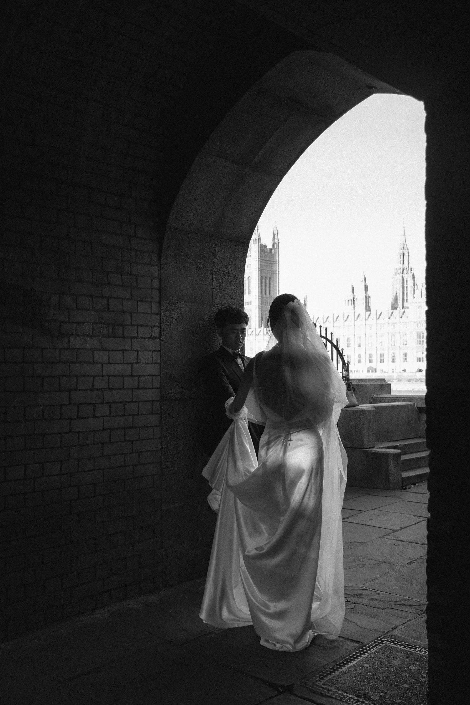 Black and white wedding photo of bride and groom under stone arch with historic building backdrop.