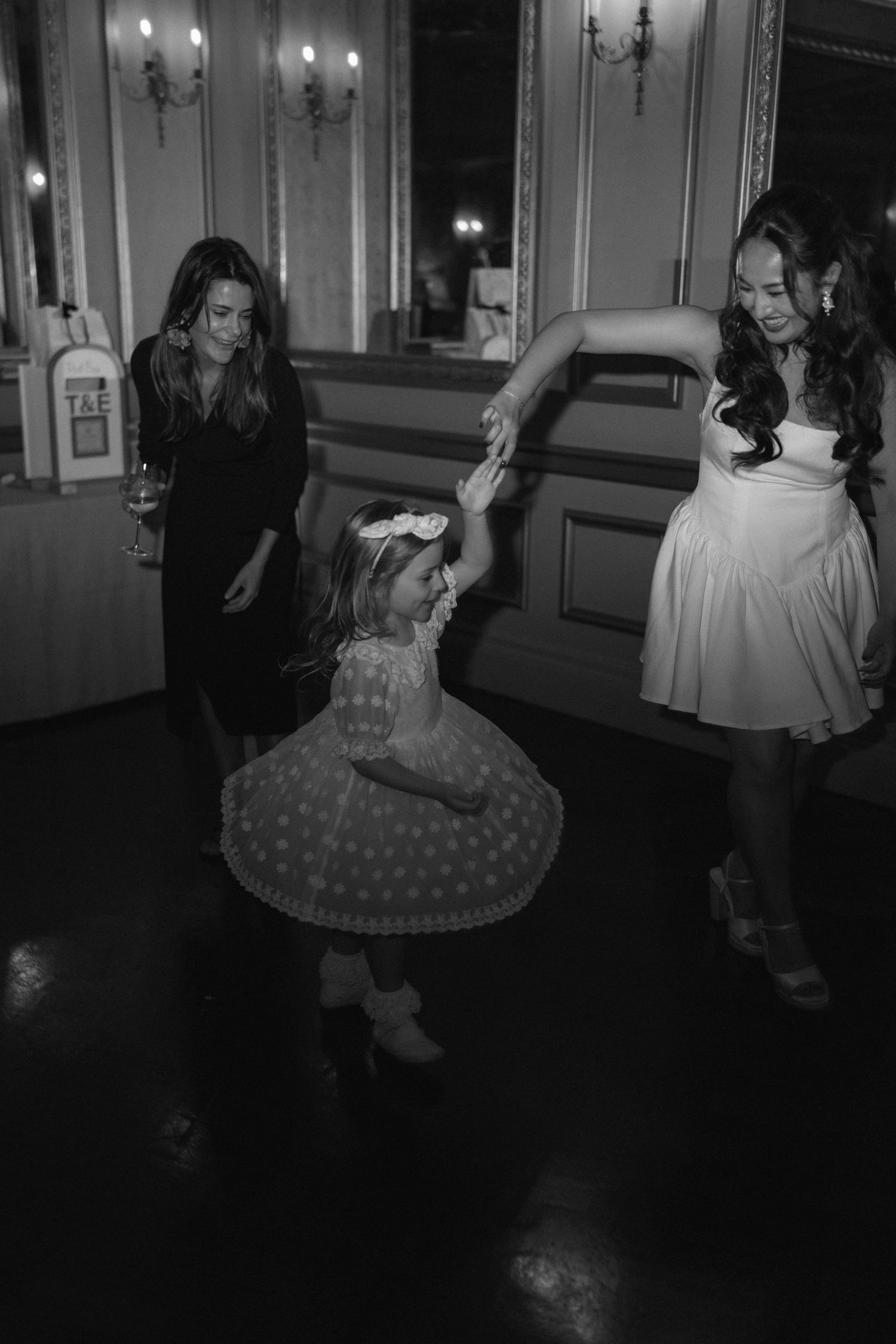 Black and white photo of a young girl dancing with a woman in a white dress at an elegant wedding reception.