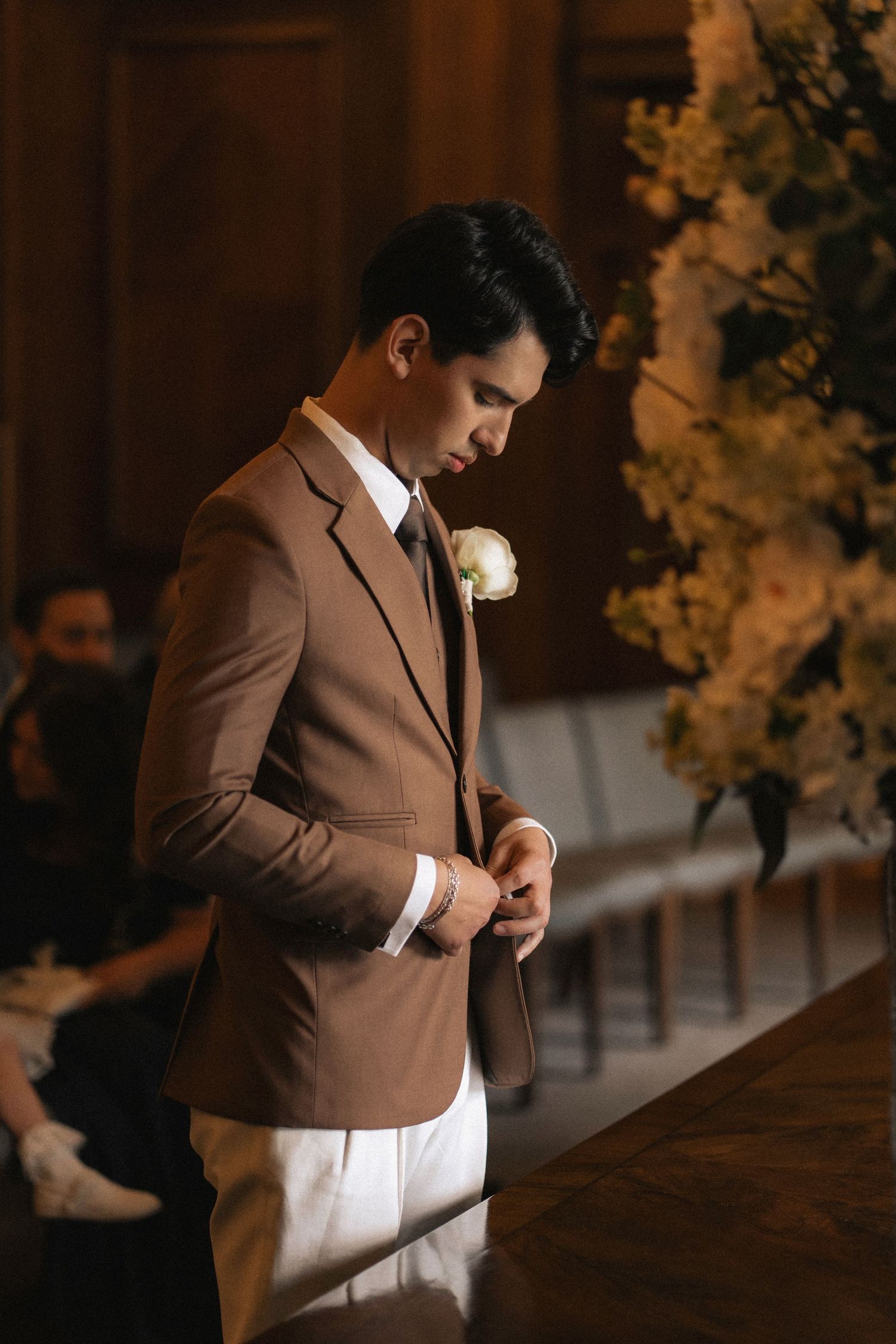 Groom in brown suit adjusts his tie while getting ready for wedding ceremony in warm lighting.