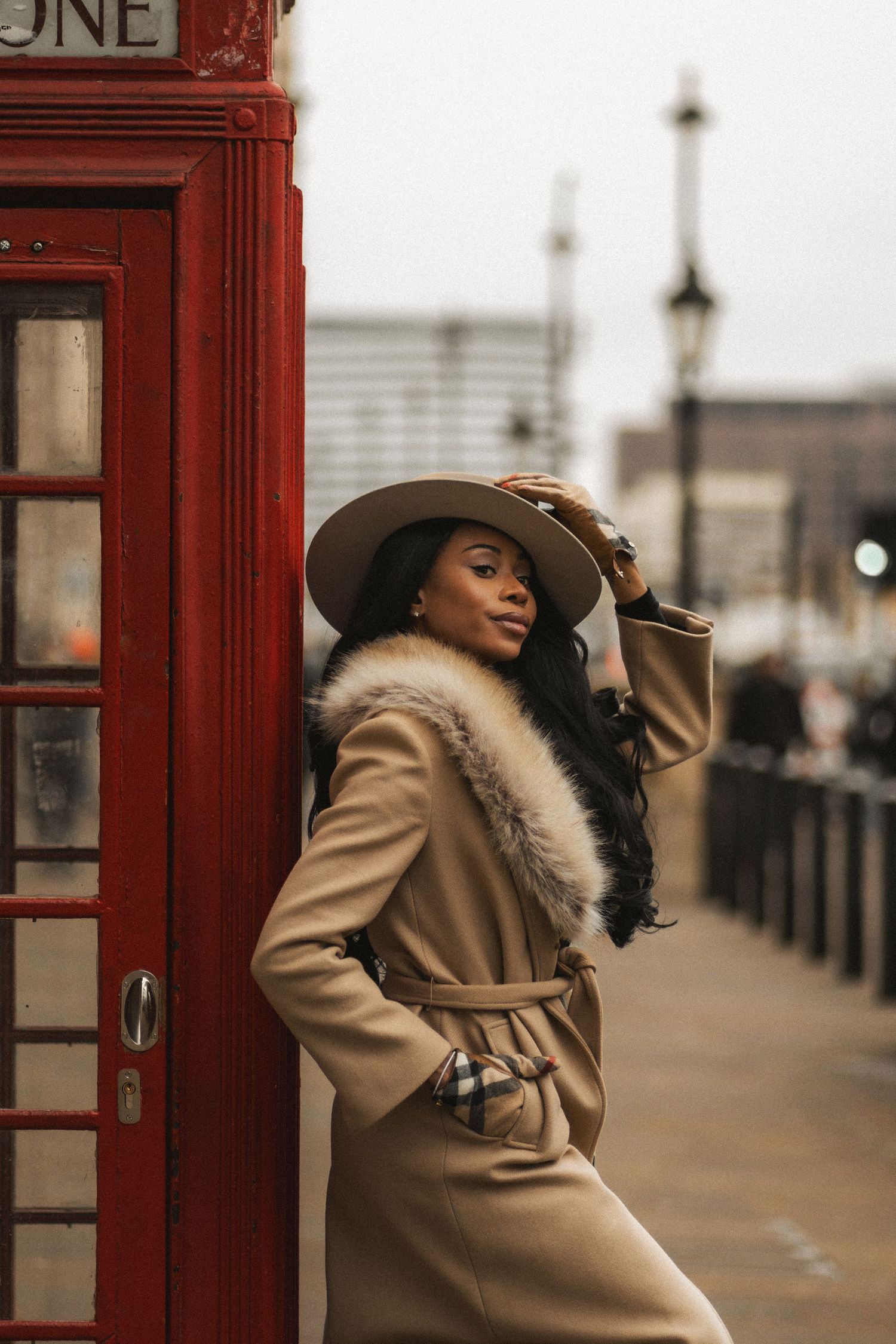 A person in a beige coat and wide-brimmed hat poses elegantly against iconic red British phone booths in London.