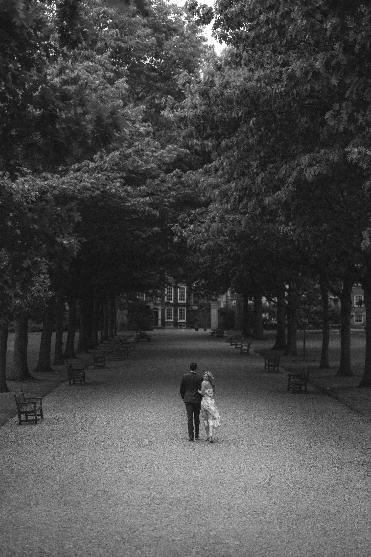 A couple walks together down a tree-lined path with benches in a peaceful park setting, captured in black and white.