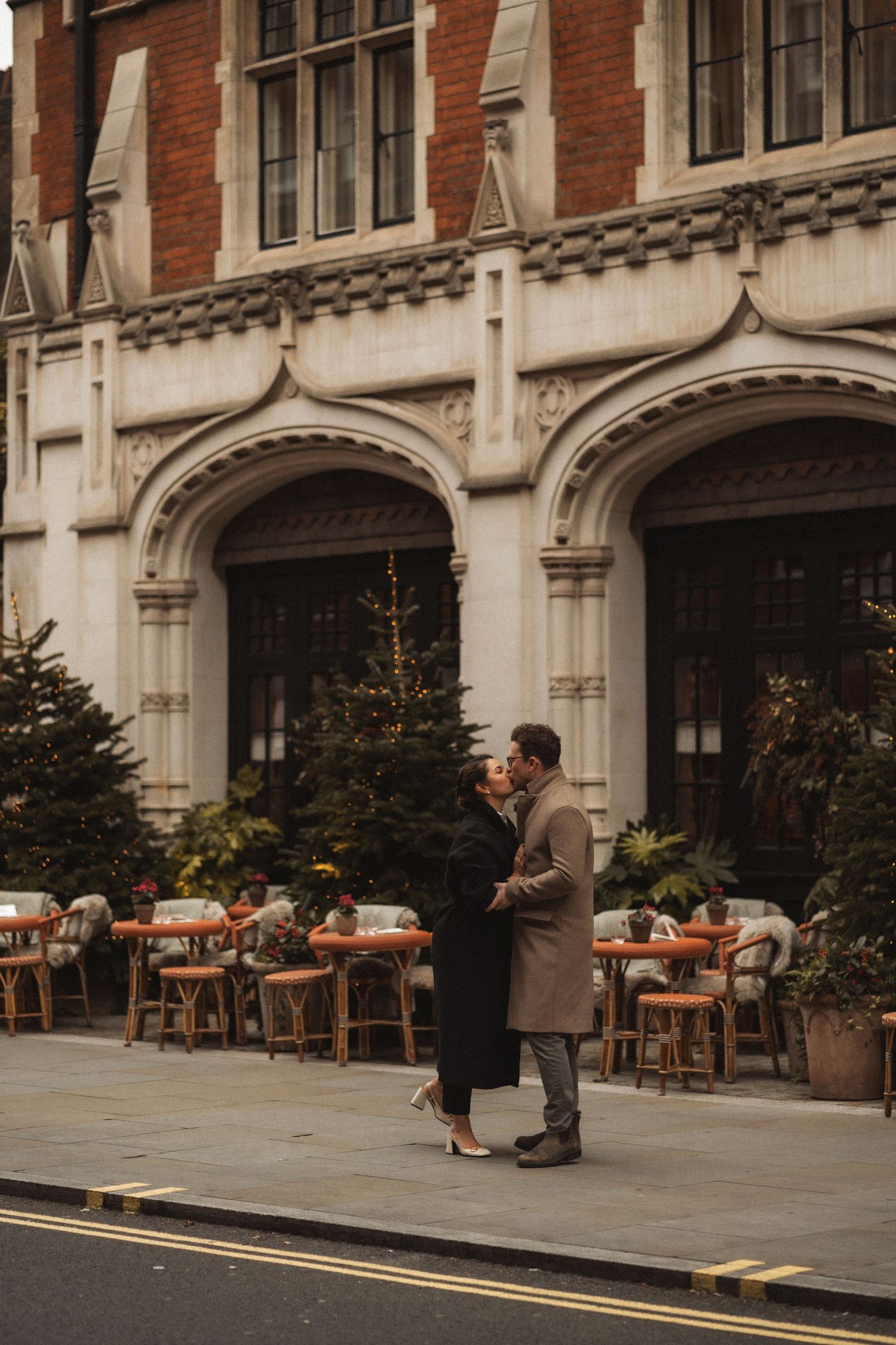 A couple stands in front of a historic brick building with outdoor restaurant seating and arched windows.