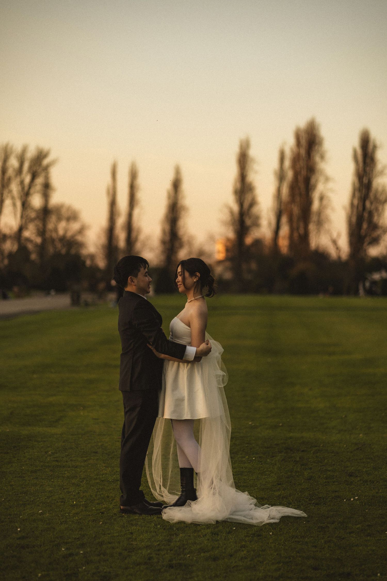 Bride and groom embrace at golden hour sunset on green lawn with tall trees in background.
