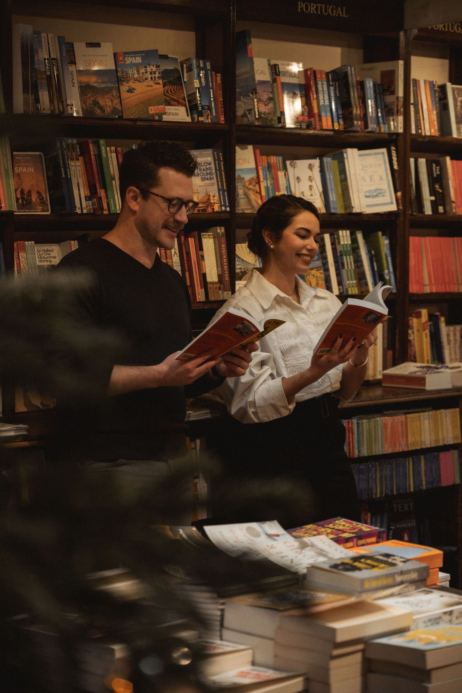 Two people browse books together at a cozy bookstore with wooden shelves in warm lighting.