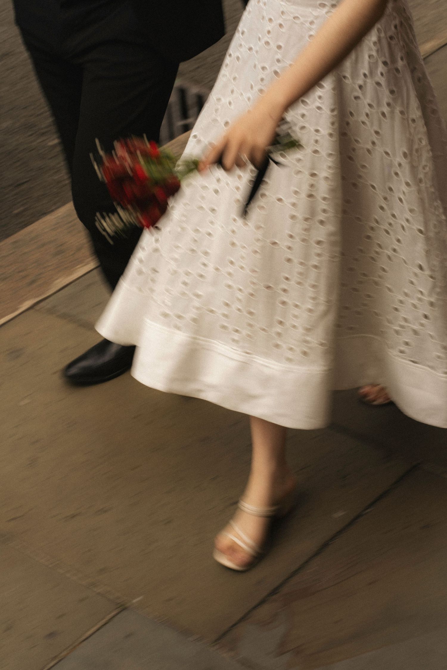 A close-up view of white eyelet lace dress fabric and bare feet walking across a wooden floor.