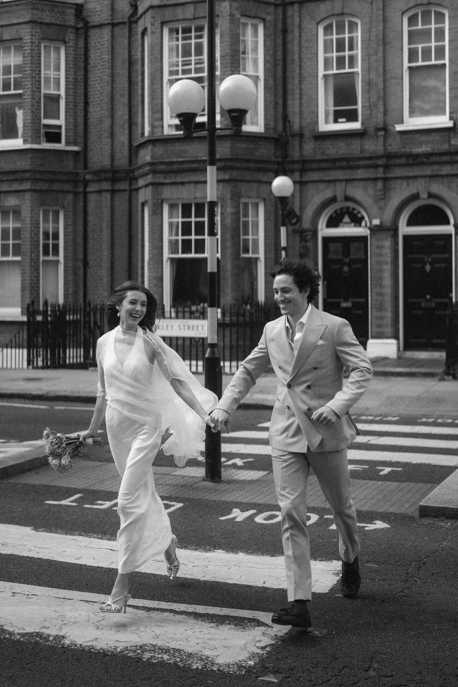 Bride and groom crossing a zebra crossing in London, holding hands and laughing in black and white photo.