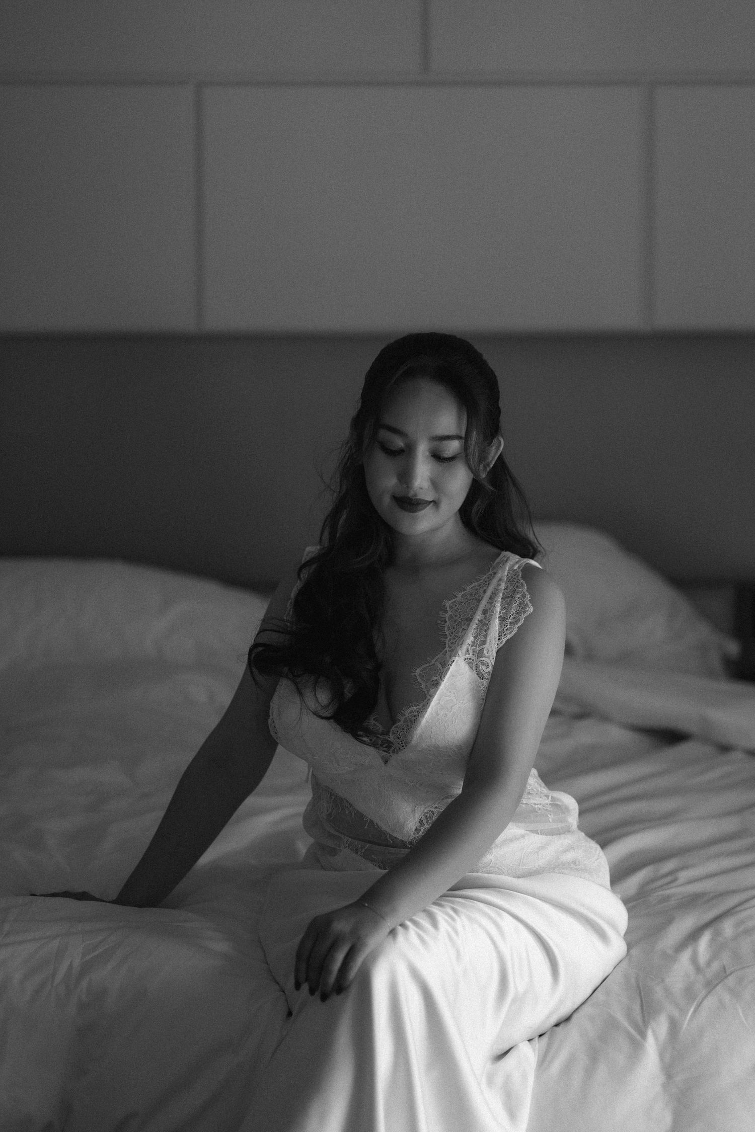 Black and white photo of a woman in a white gown sitting on a bed, looking down with elegant poise.