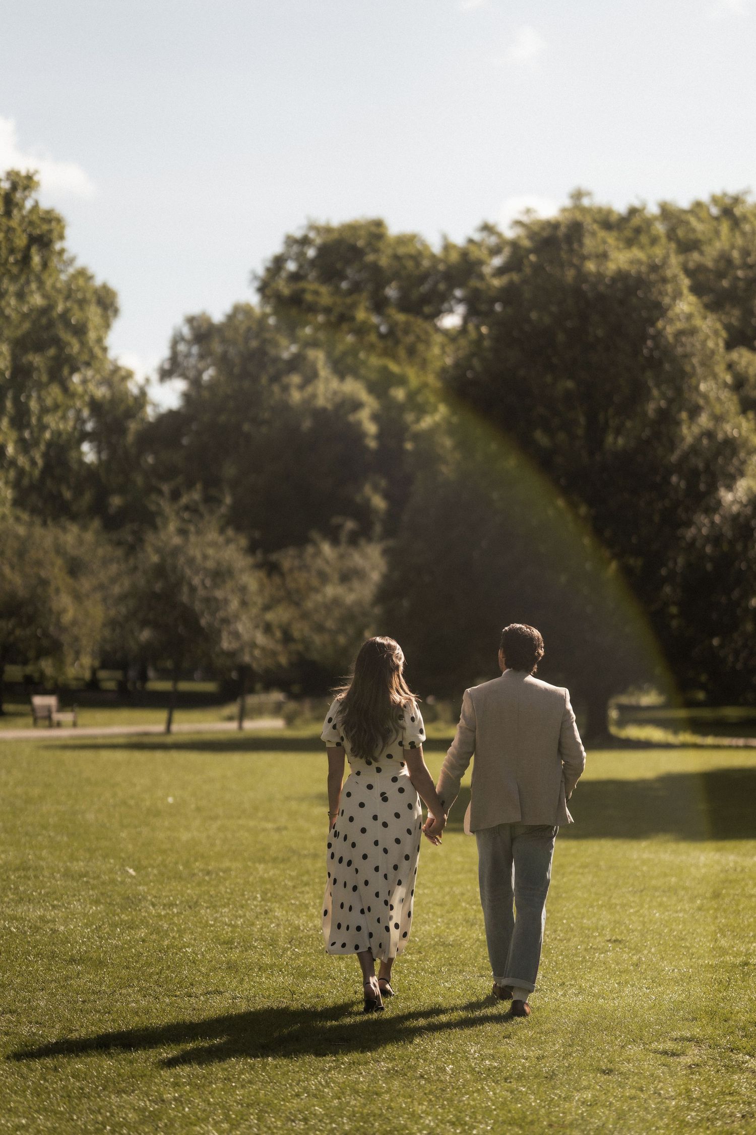 Couple holding hands walking through a sunny green park, woman in polka dot dress and man in beige suit.