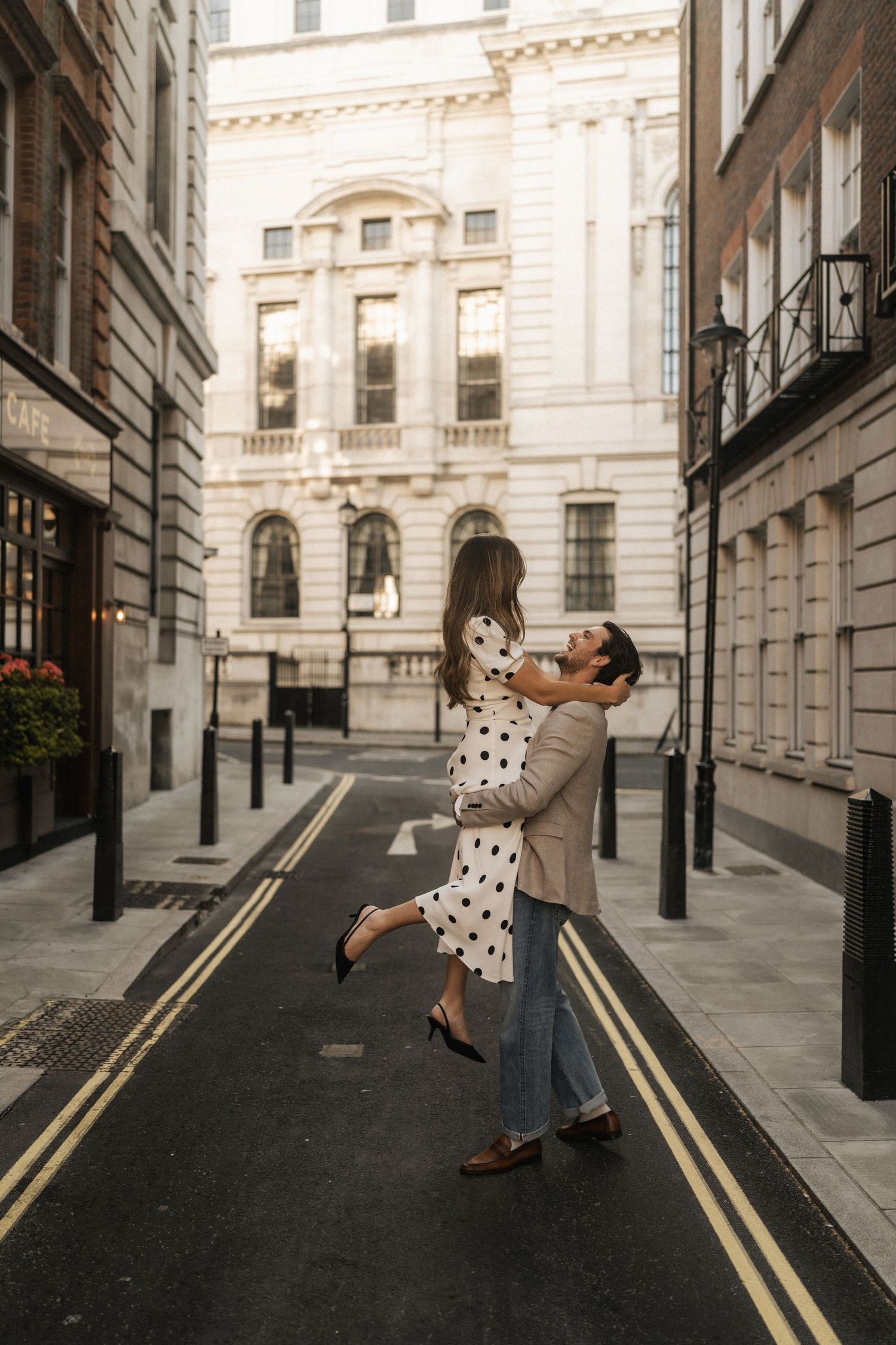 Couple dancing romantically on a quiet London street, woman in polka dot dress lifted by man in beige blazer.