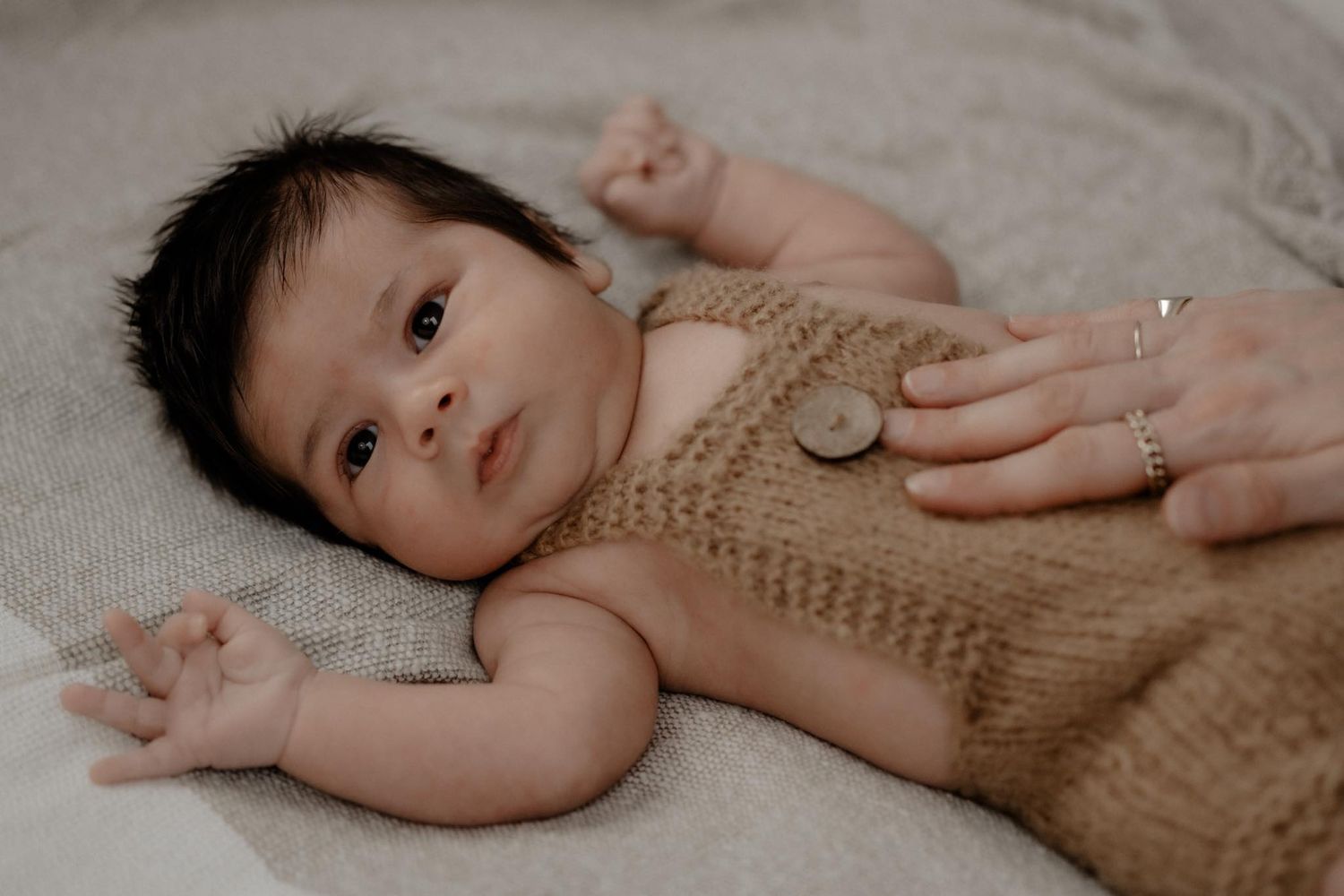 A baby wearing a tan knit romper lays on a light gray blanket while an adult's hand gently touches their chest.