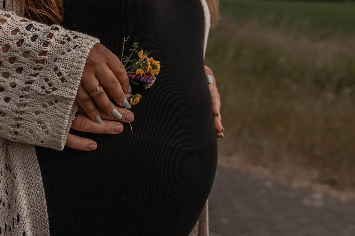 A pregnant belly silhouetted against sunlight while hands hold small yellow wildflowers in a knitted sweater sleeve.