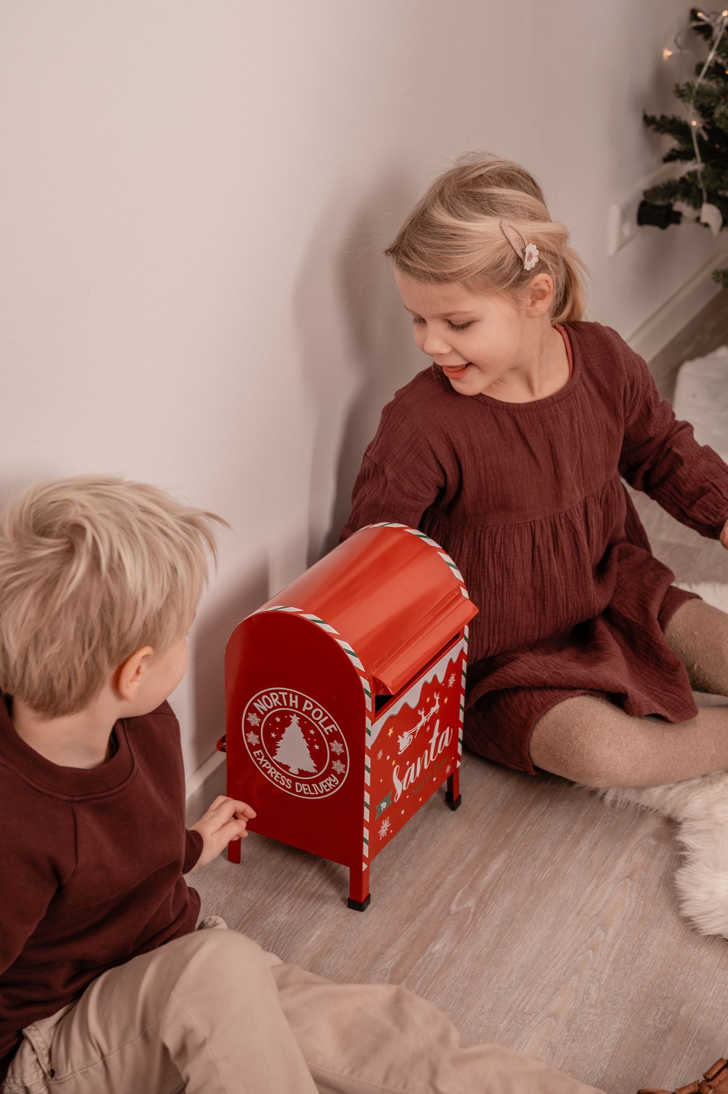 Children in burgundy outfits playing with a festive red Santa's mailbox on a wooden floor.