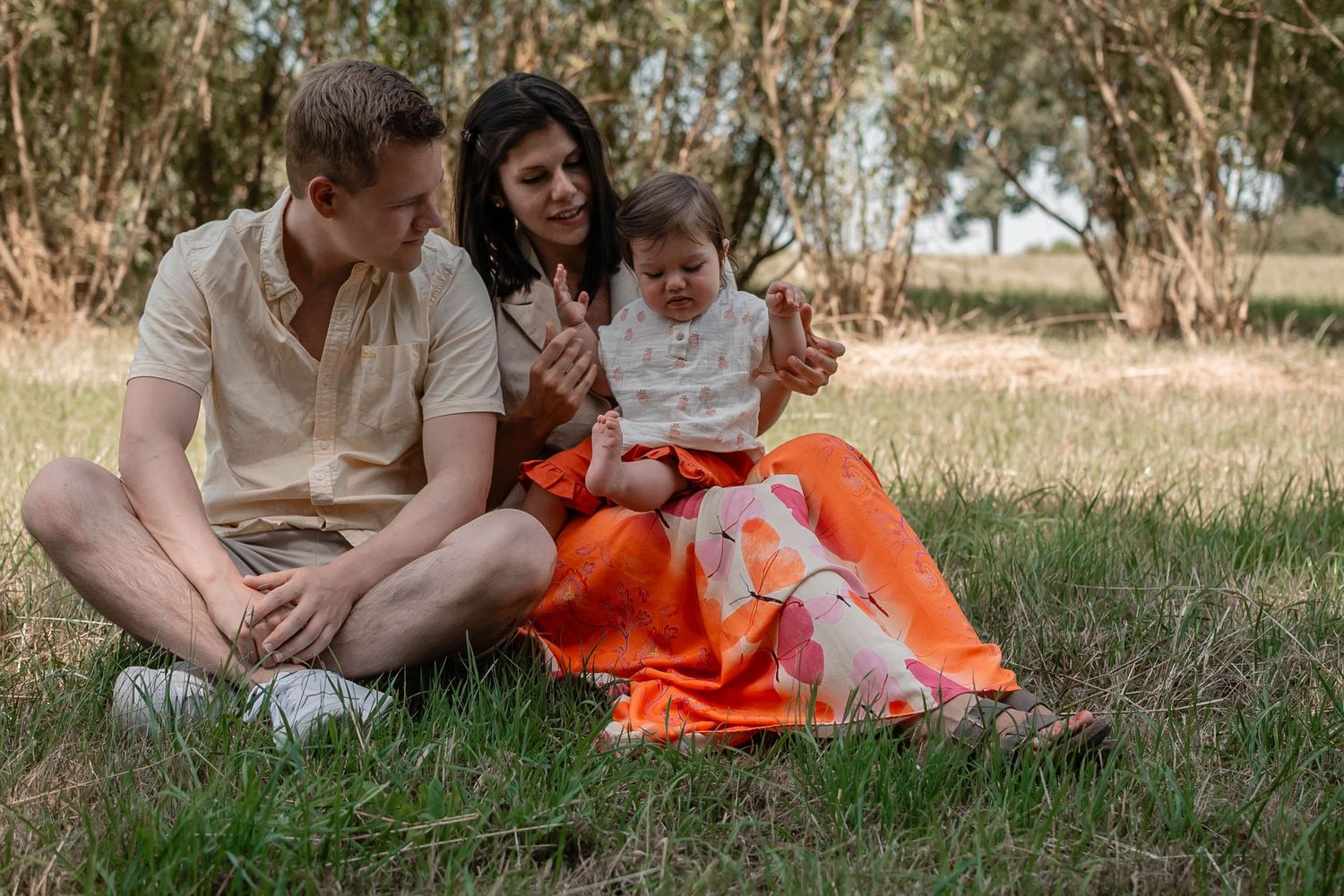 A family sits together on an orange blanket having a picnic in a grassy area surrounded by trees on a sunny day.