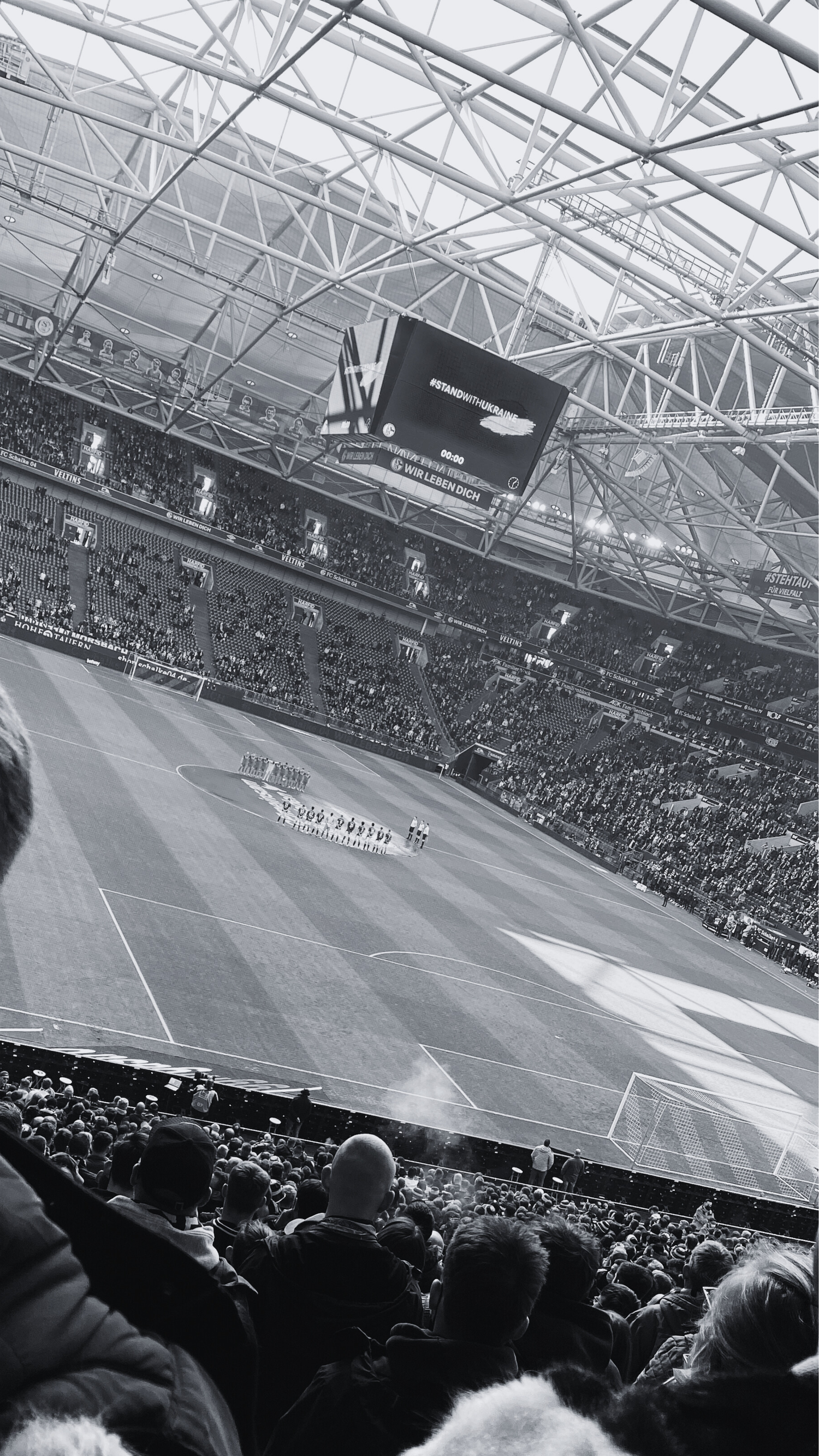 Black and white view of a packed soccer stadium with large scoreboard and crowd watching teams line up on the field.