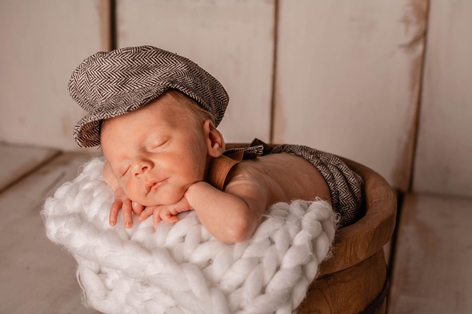 Sleeping newborn wearing grey flat cap rests peacefully on chunky knit white blanket during professional photo session.