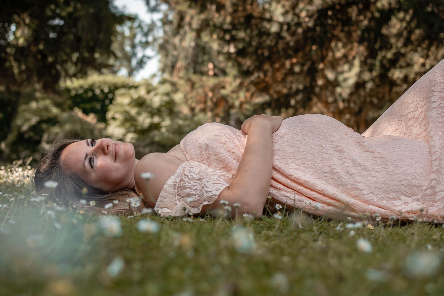 A person in a blush pink lace dress lies peacefully in sunlit grass during an outdoor photo session.