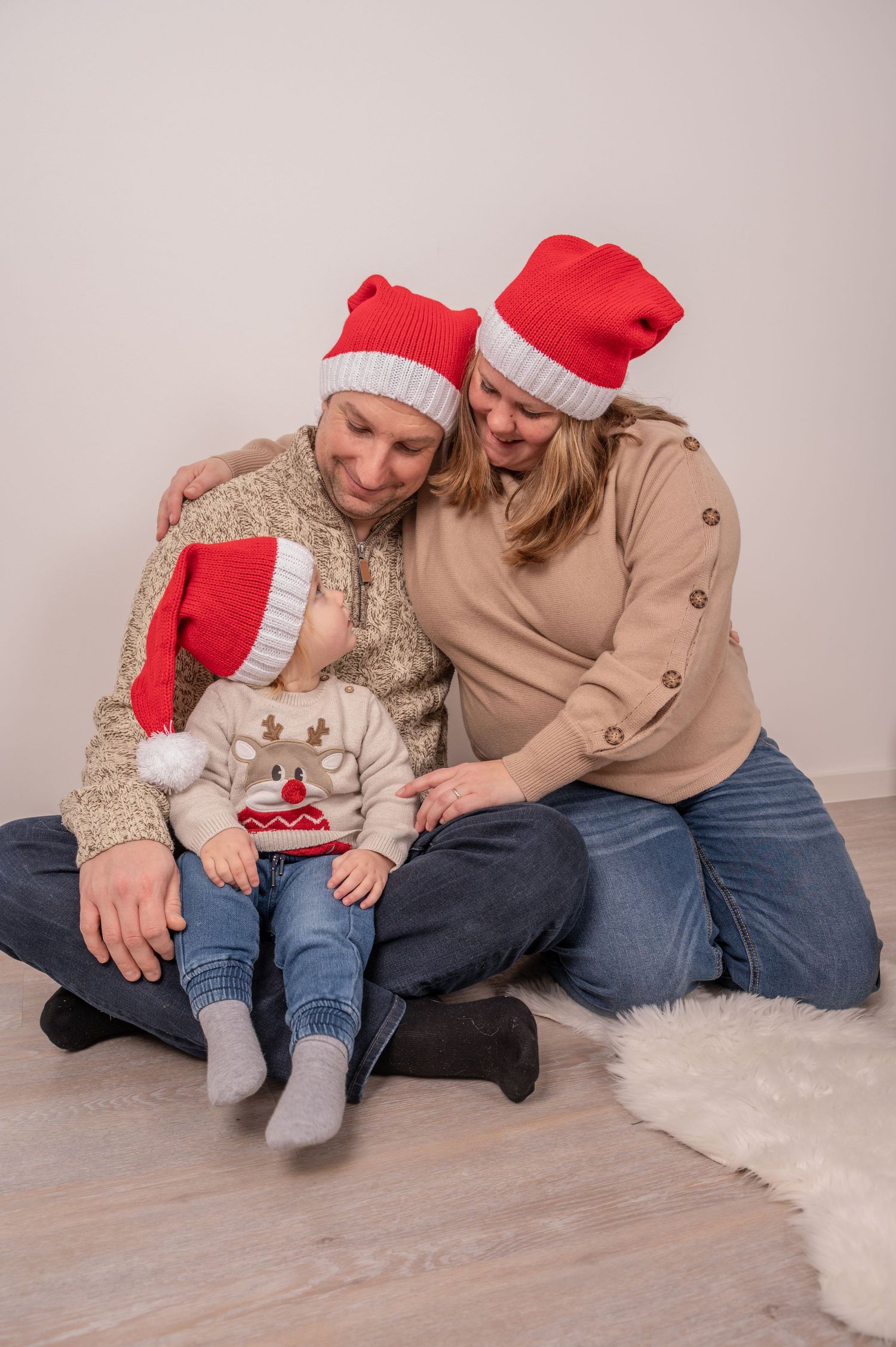 Two people wearing Santa hats share a tender moment on a white fluffy rug in matching holiday attire.
