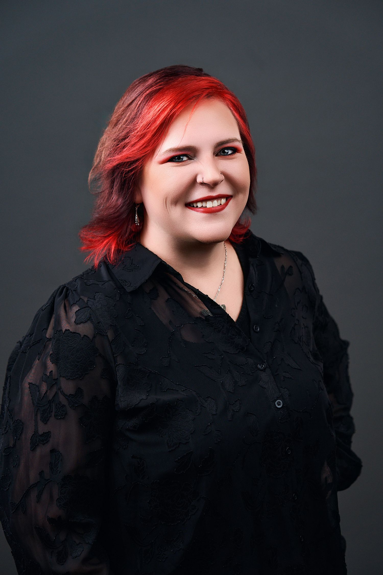 Smiling woman with vibrant red hair wearing a black lace blouse against a dark gray studio background.