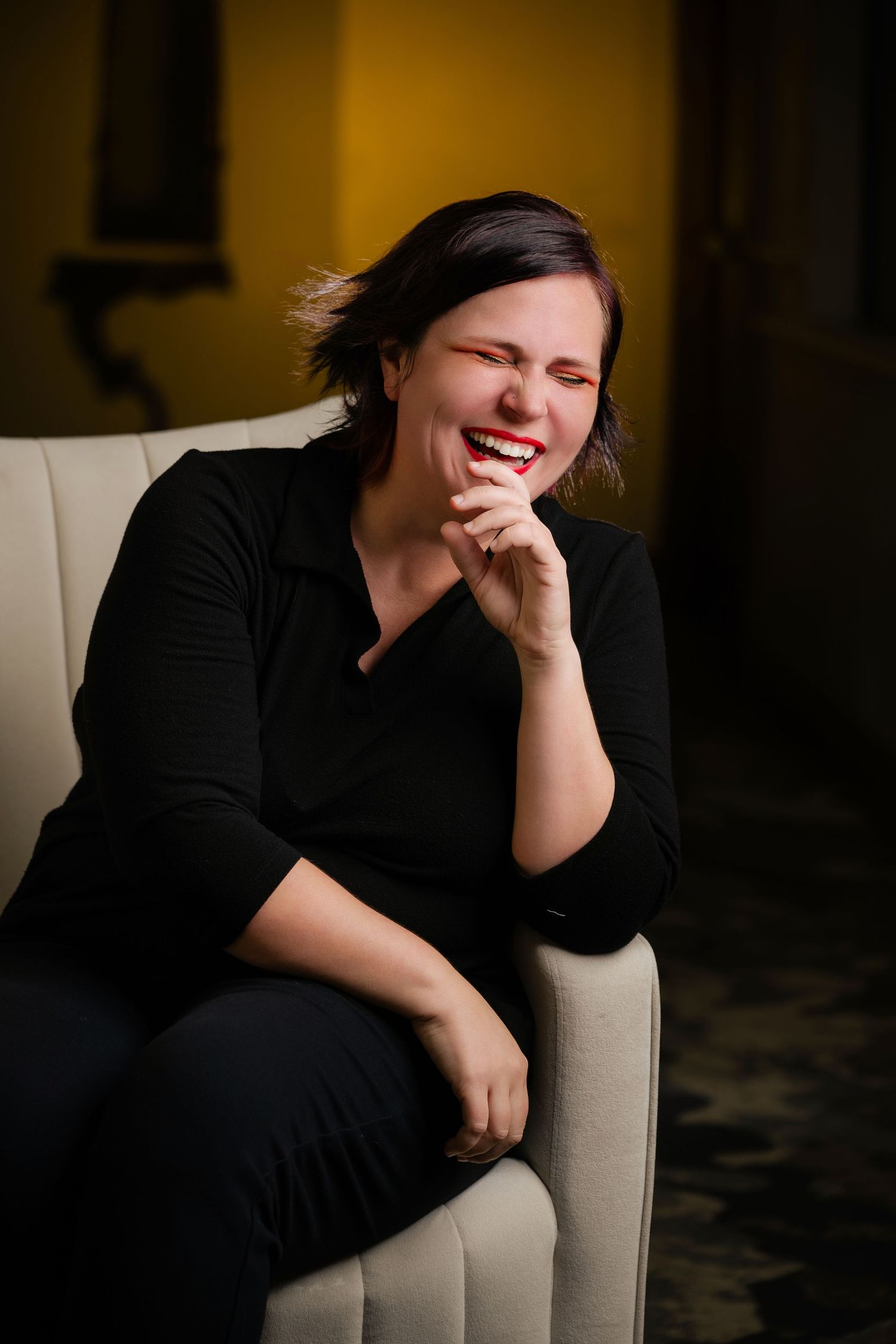 Daniella Koontz wearing a black sweater sits comfortably in a cream-colored chair while laughing joyfully in warm lighting.