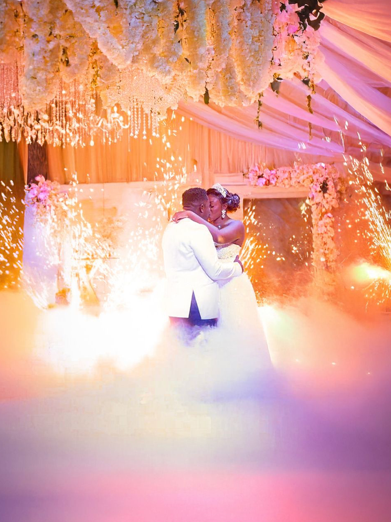 A romantic wedding reception scene with sparklers creating a magical atmosphere under elegant crystal chandeliers and pink lighting.