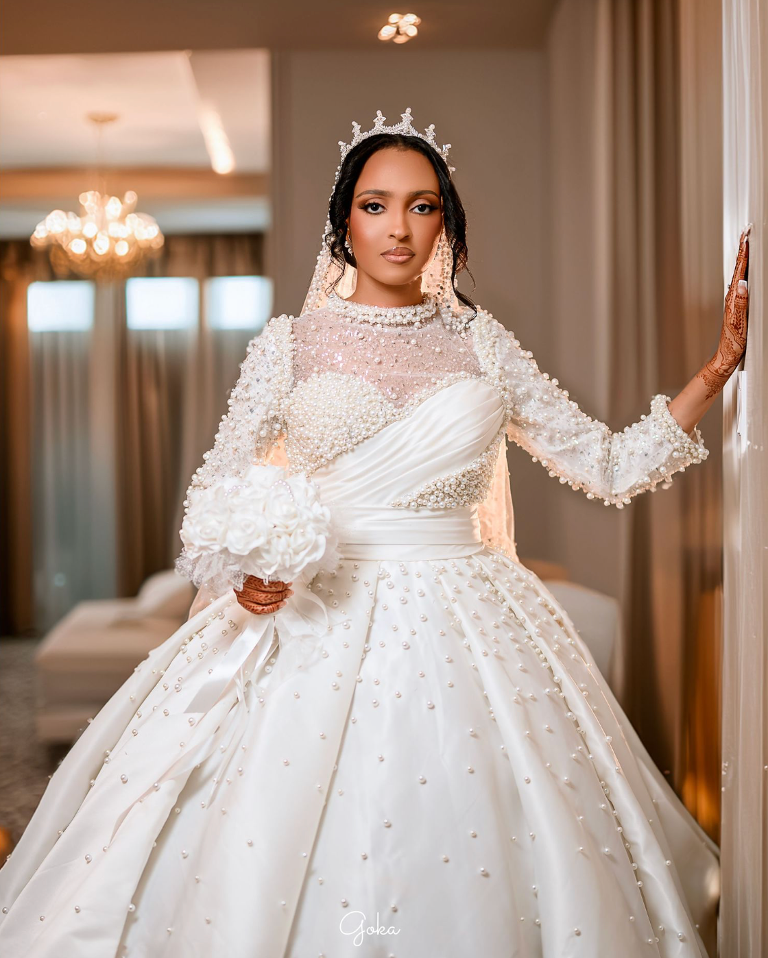 Elegant bride in a luxurious white ballgown with pearls and lace details poses in a warmly lit hallway.