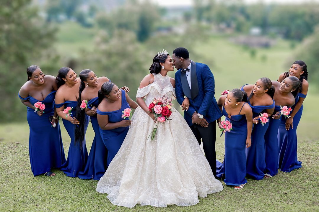 A row of bridesmaids in royal blue dresses sit on grass while the bride in white leans over them in a candid moment.