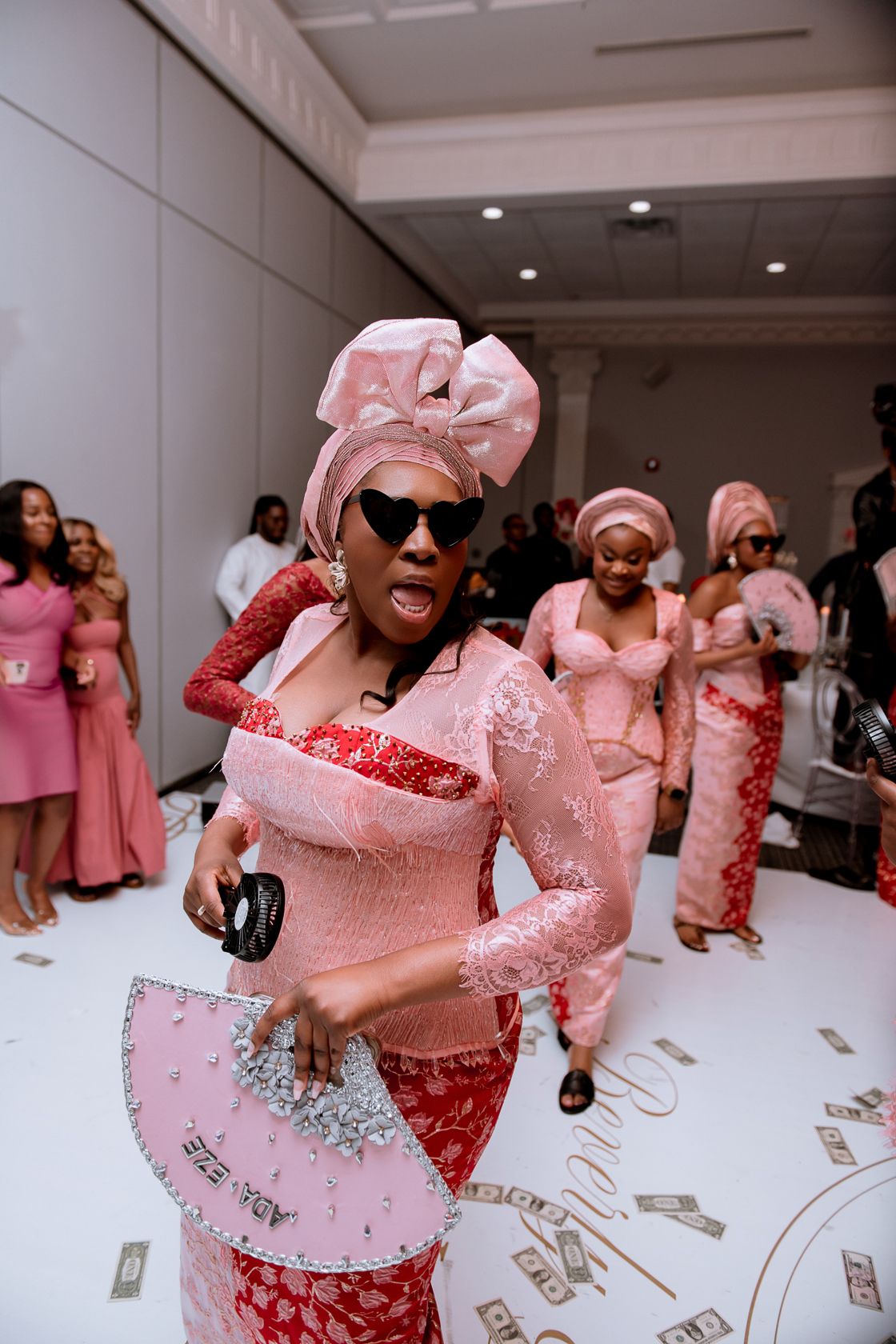 Joyful partygoers in pink outfits dance and celebrate as money rains down around them at an indoor event.