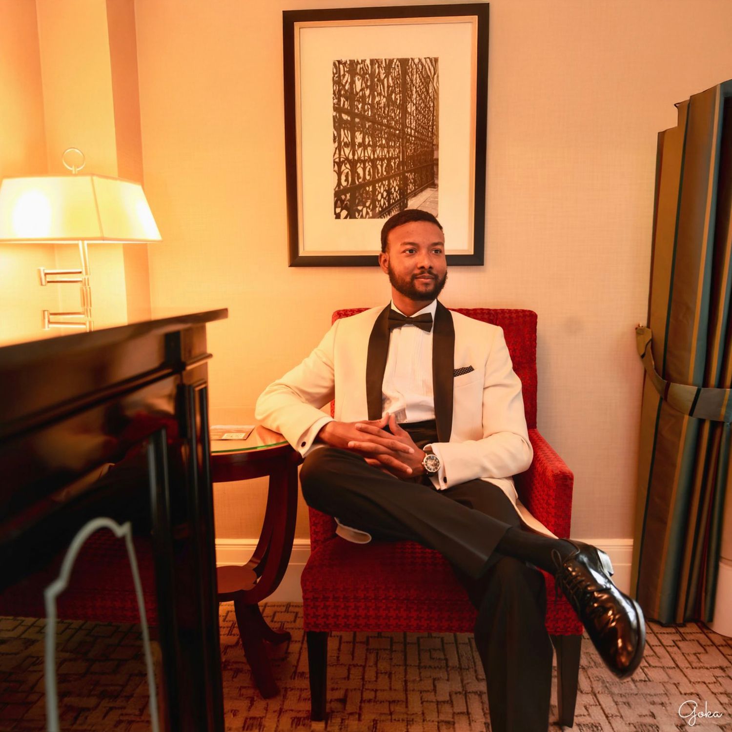 A person in a tuxedo sits on a chair in a warmly lit room with framed artwork on the wall.