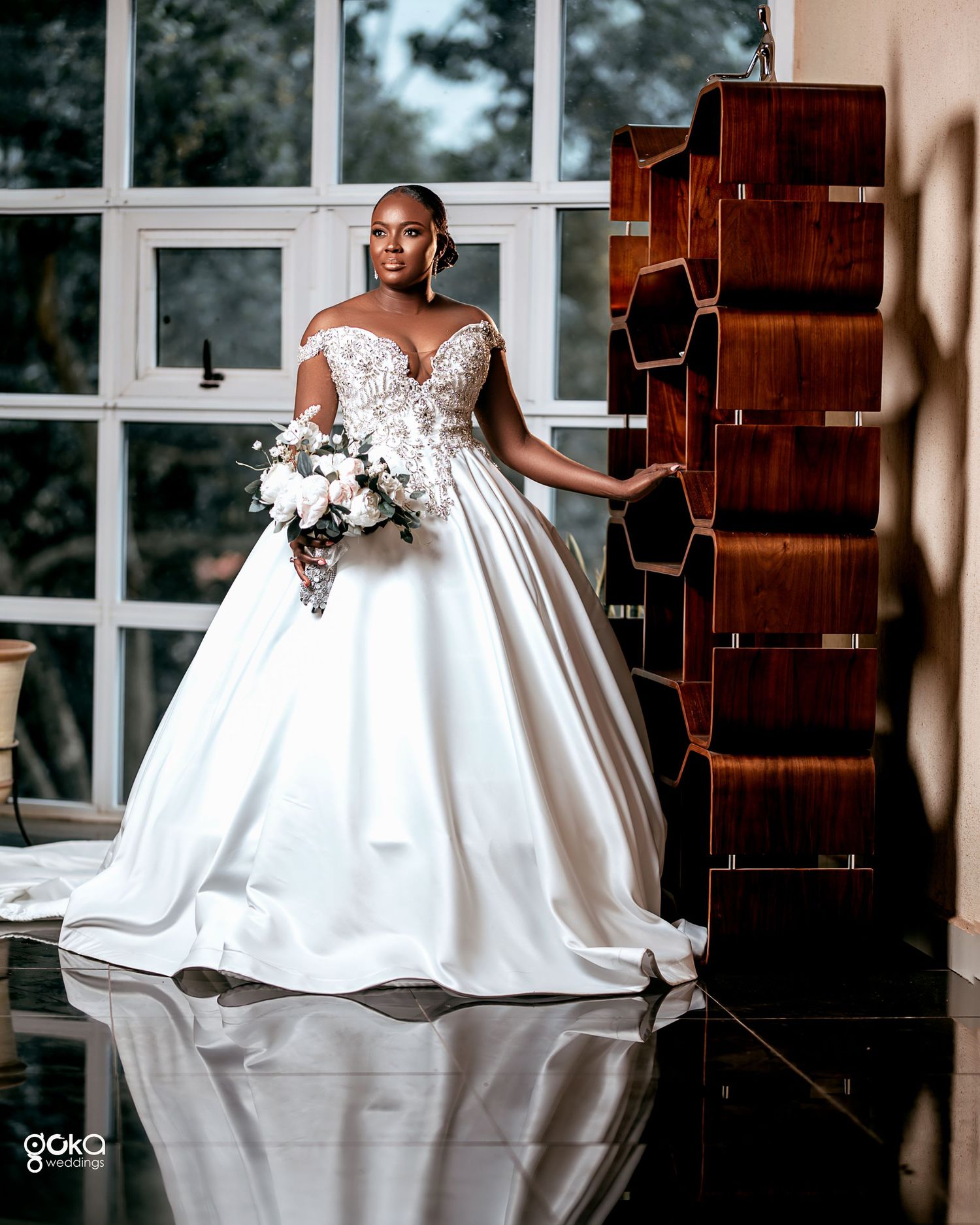 A bride in a stunning white satin ball gown poses elegantly in a sunlit room with wooden accents.