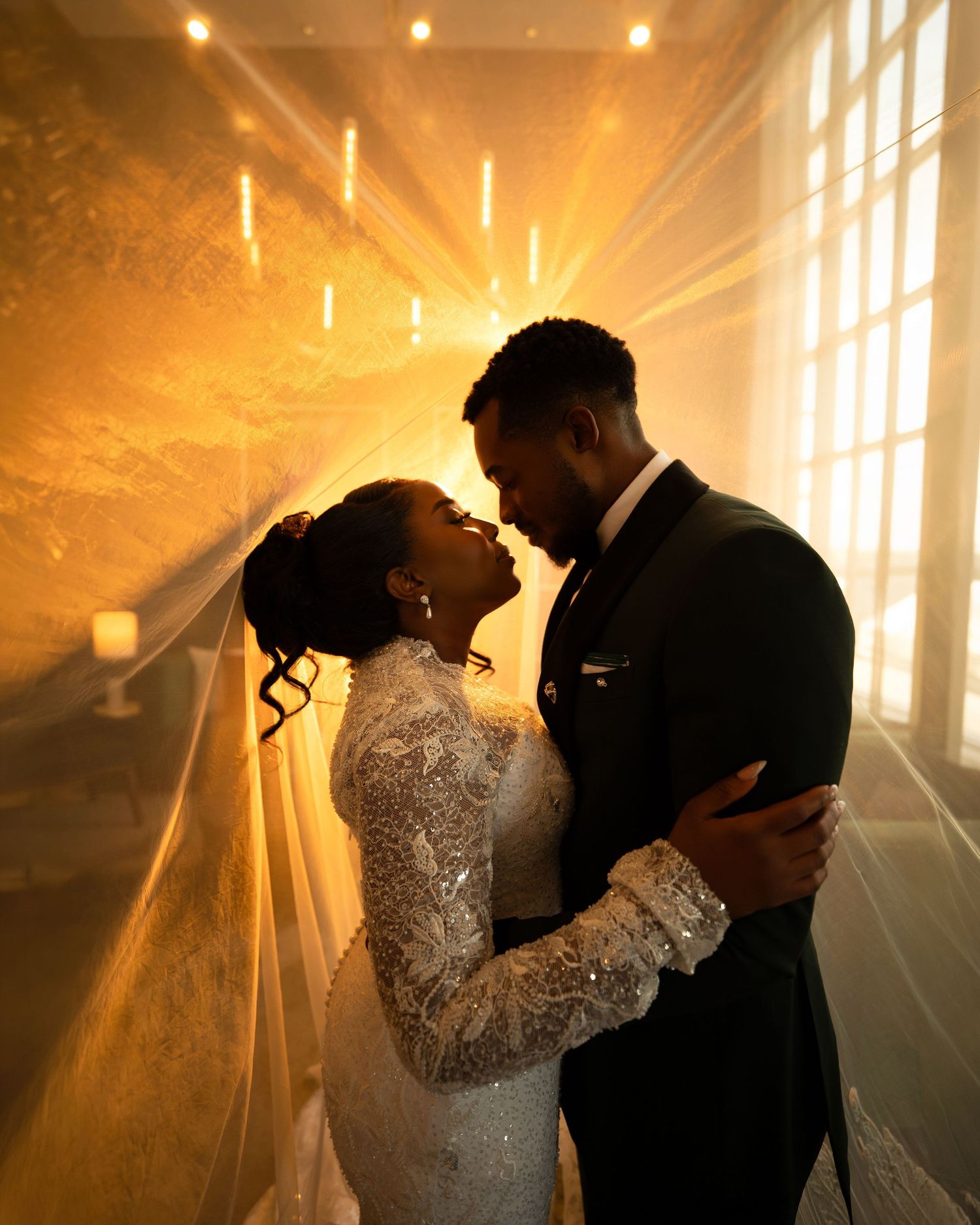 A Black bride and groom share a romantic embrace in golden light, her beaded gown and veil glowing warmly.