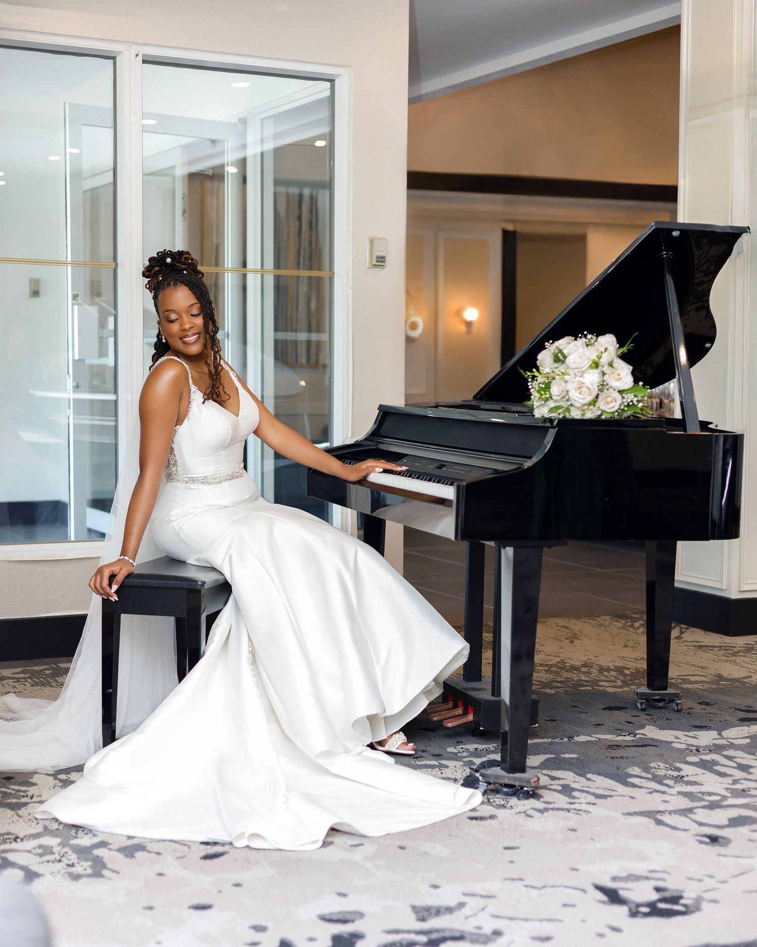 A figure in an elegant white wedding dress poses gracefully by a grand piano in an elegant room with patterned carpet.