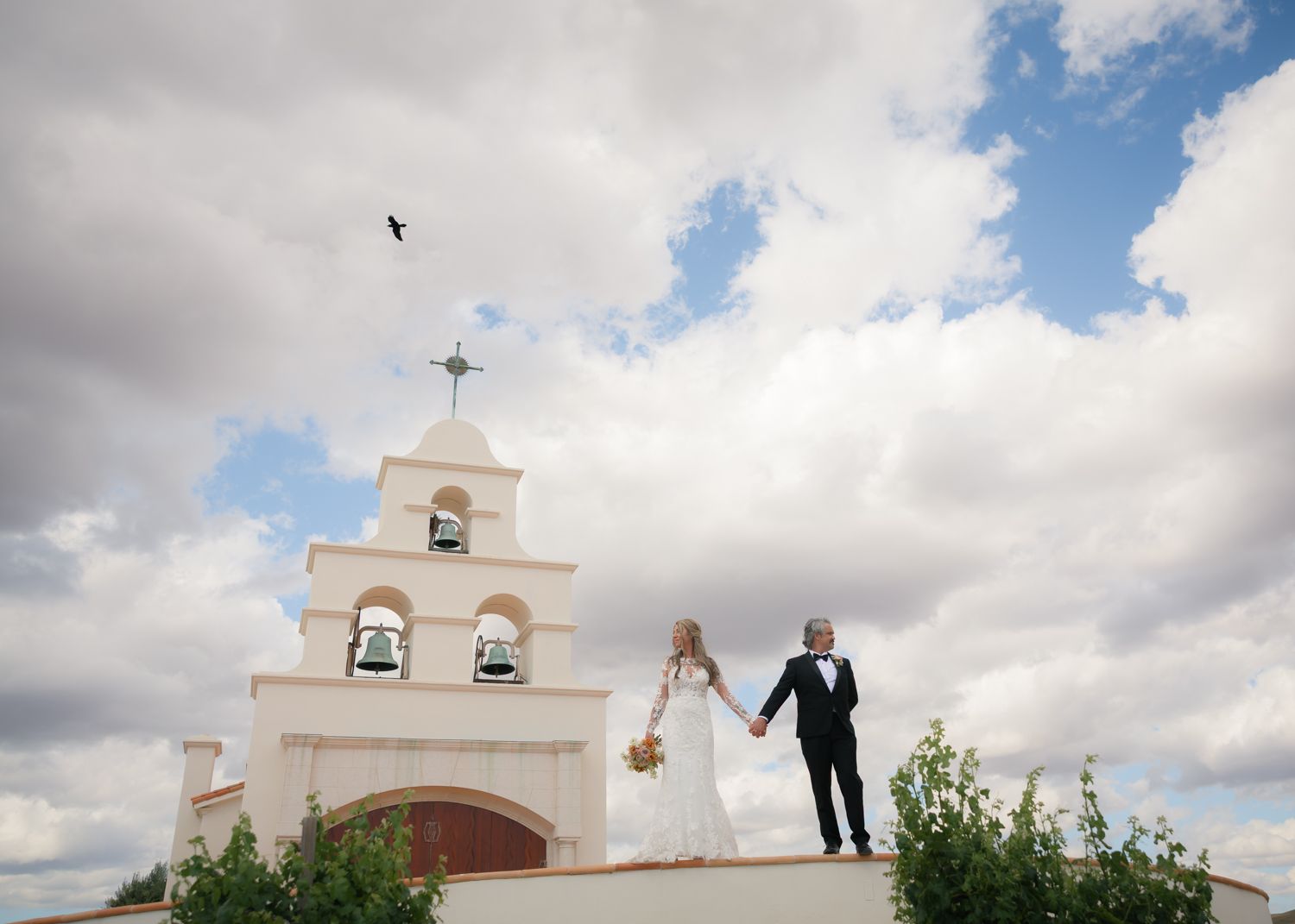 Bride and groom hold hands atop a white Spanish mission bell tower under dramatic cloudy blue skies.