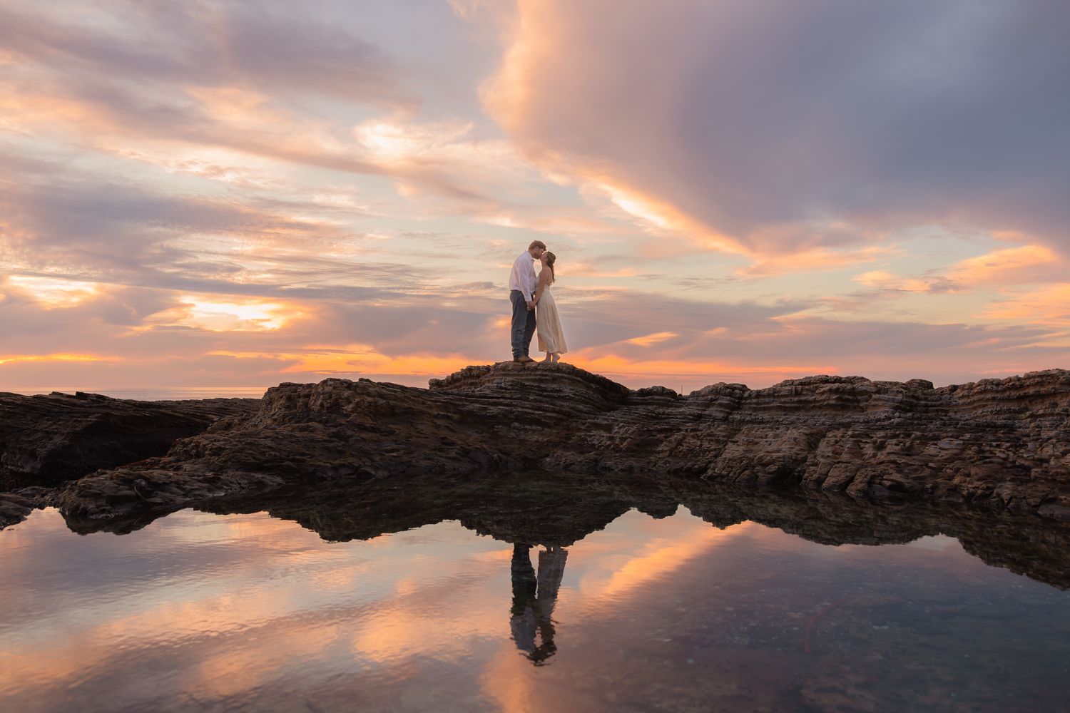 Silhouetted figures stand on rocky outcrop against dramatic sunset sky with perfect reflection in still water.