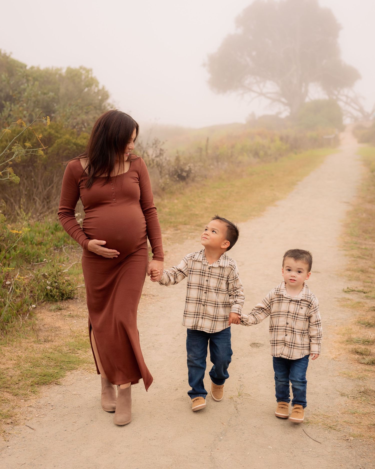 A parent and child walk together on a misty dirt path through coastal brush while holding hands.